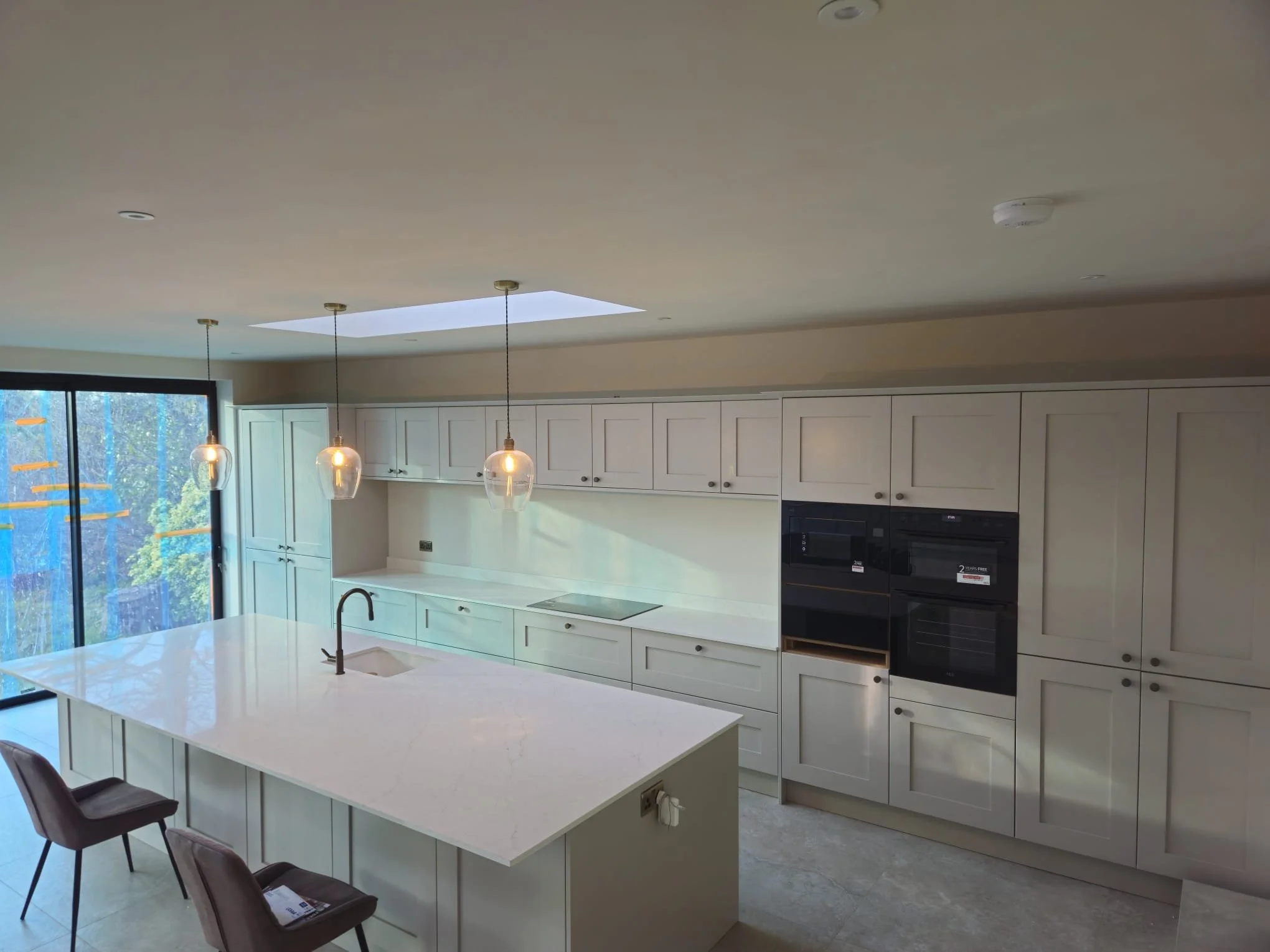 Modern kitchen with a sleek white island countertop and a dark faucet. Three pendant lights hang above. Cabinets and a large window in the background.
