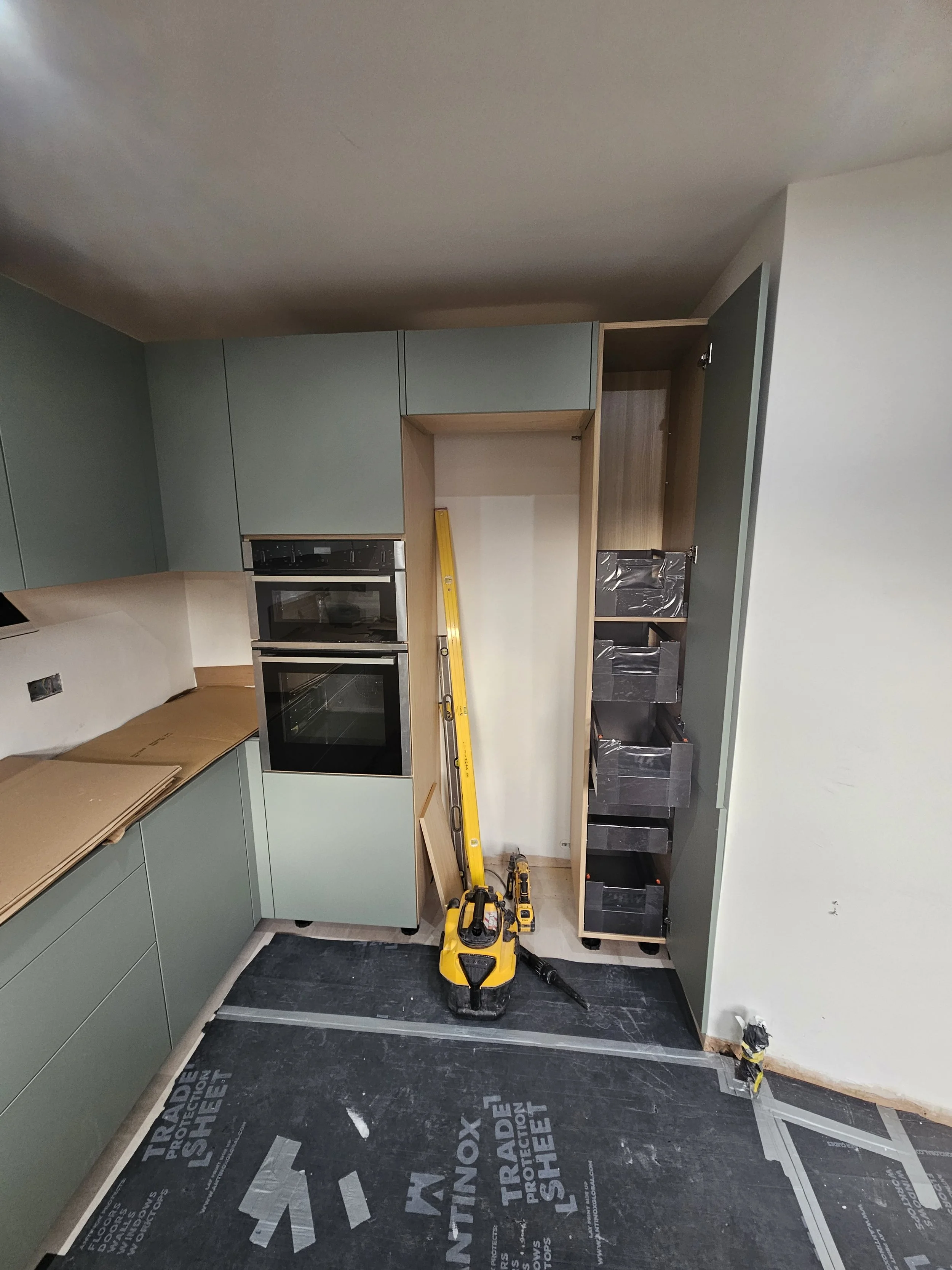 Modern kitchen under construction with mint green cabinets, double ovens, and a stack of drawers. A yellow level and protective sheets are visible.