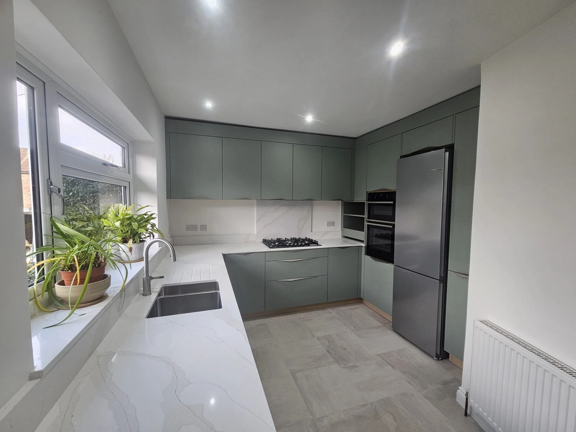 Modern kitchen with green cabinets, marble countertops, and stainless steel appliances. A window with plants adds a fresh touch, under bright ceiling lights.