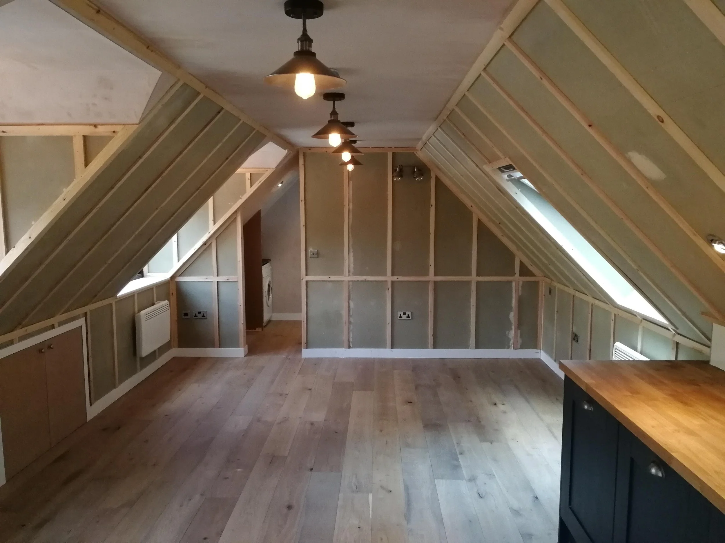 An attic room with wooden paneling and sloped ceilings, featuring exposed pendant lights. Light wood flooring adds a warm and cozy feel.