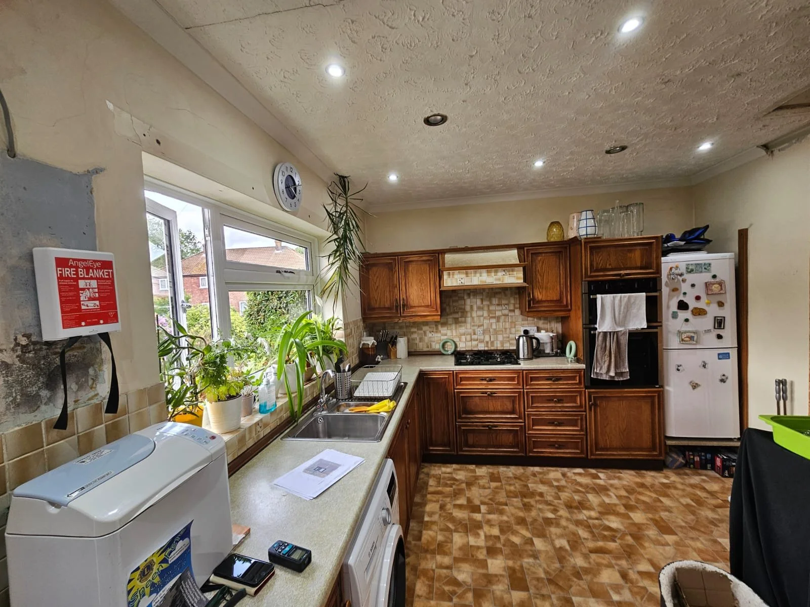 A cozy kitchen with wooden cabinets and tiled backsplash. A window above the sink lets in natural light, illuminating plants on the sill. The atmosphere is warm and inviting.