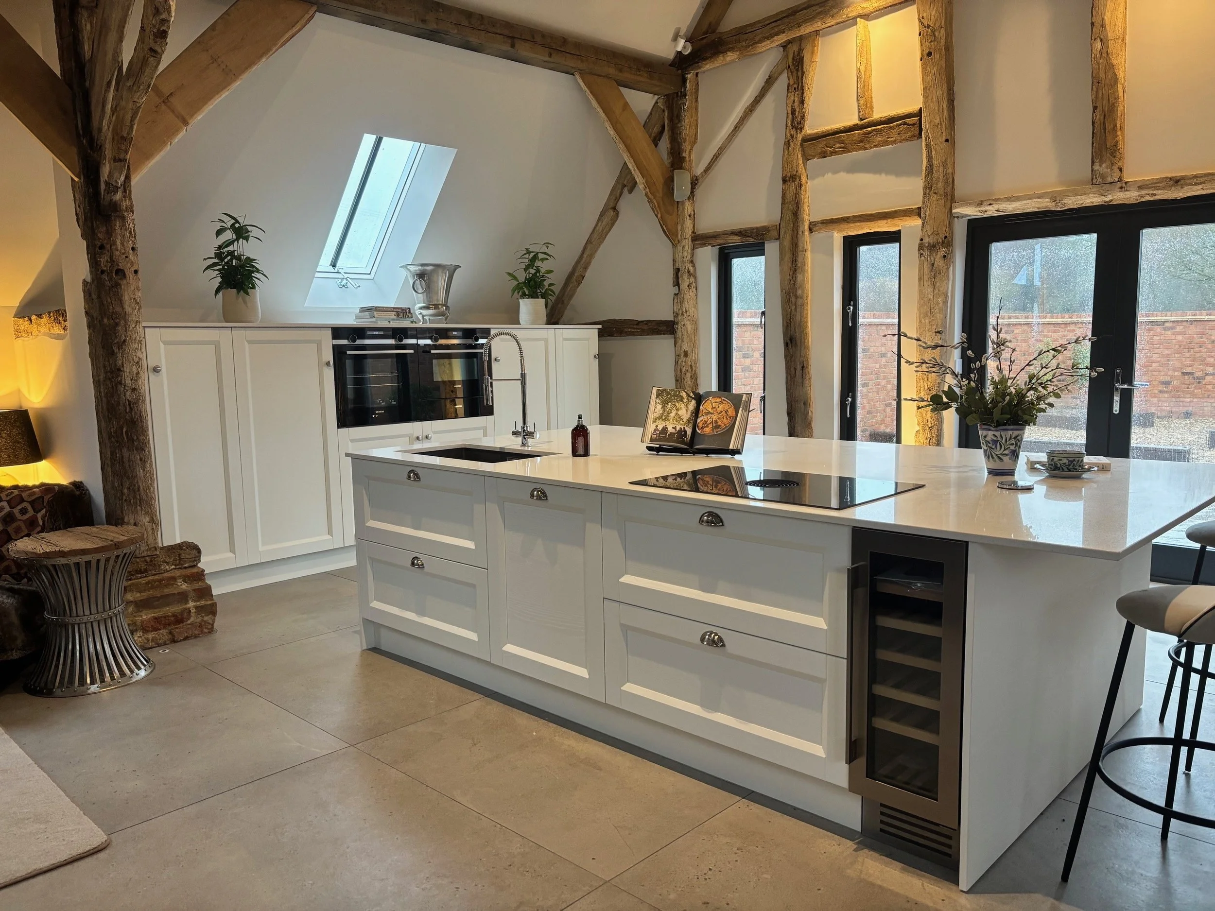 Modern rustic kitchen with a white island, integrated oven, and wooden beams. Soft lighting creates a warm, inviting atmosphere. Skylight and potted plants add freshness.