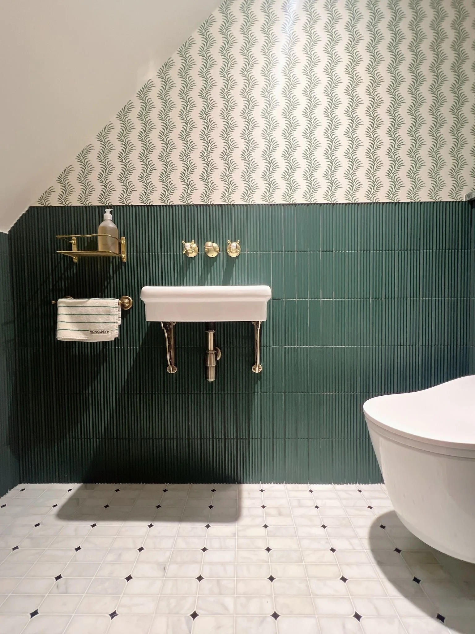 A small bathroom features a geometric wavy-patterned wallpaper above dark green tiles. It includes a wall-mounted sink with brass fixtures. Modern and stylish.