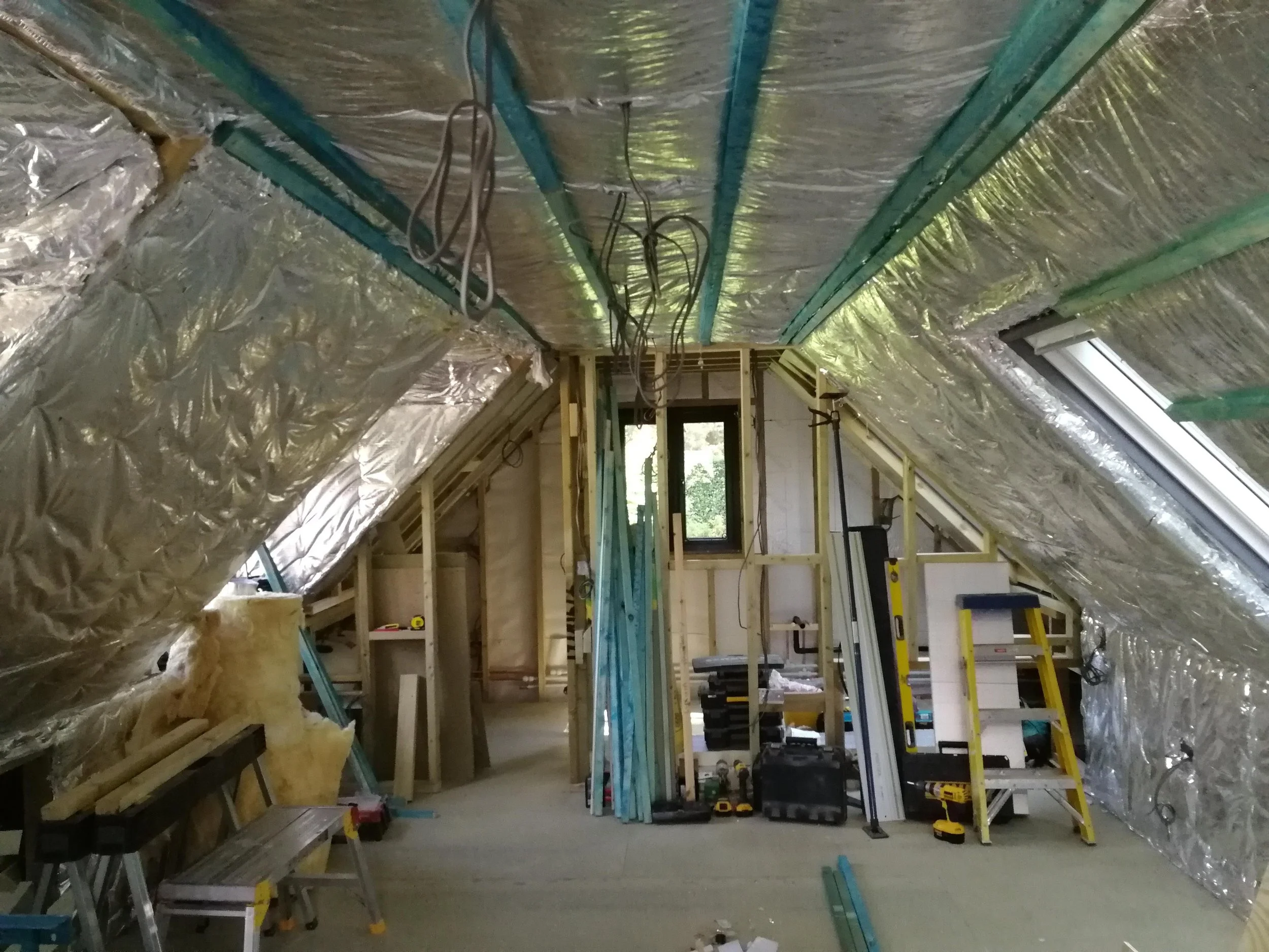 An attic under renovation with exposed insulation and wooden framing, scattered tools, and a stepladder, conveying a work-in-progress atmosphere.