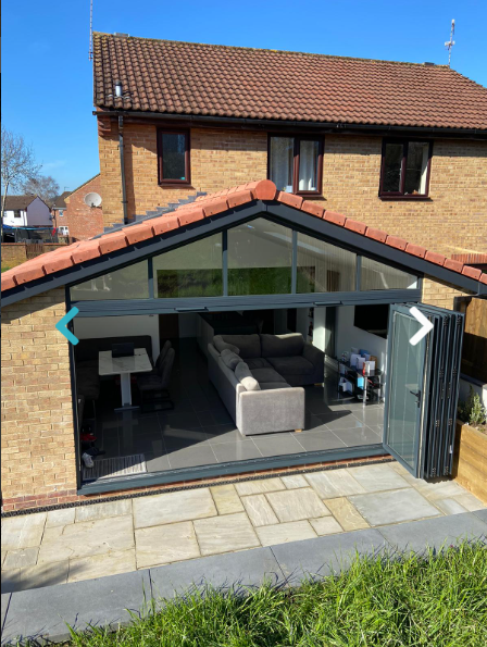 A brick house with a modern glass extension featuring a large open door. Inside, a cozy living space with a sectional sofa and a dining area is visible.
