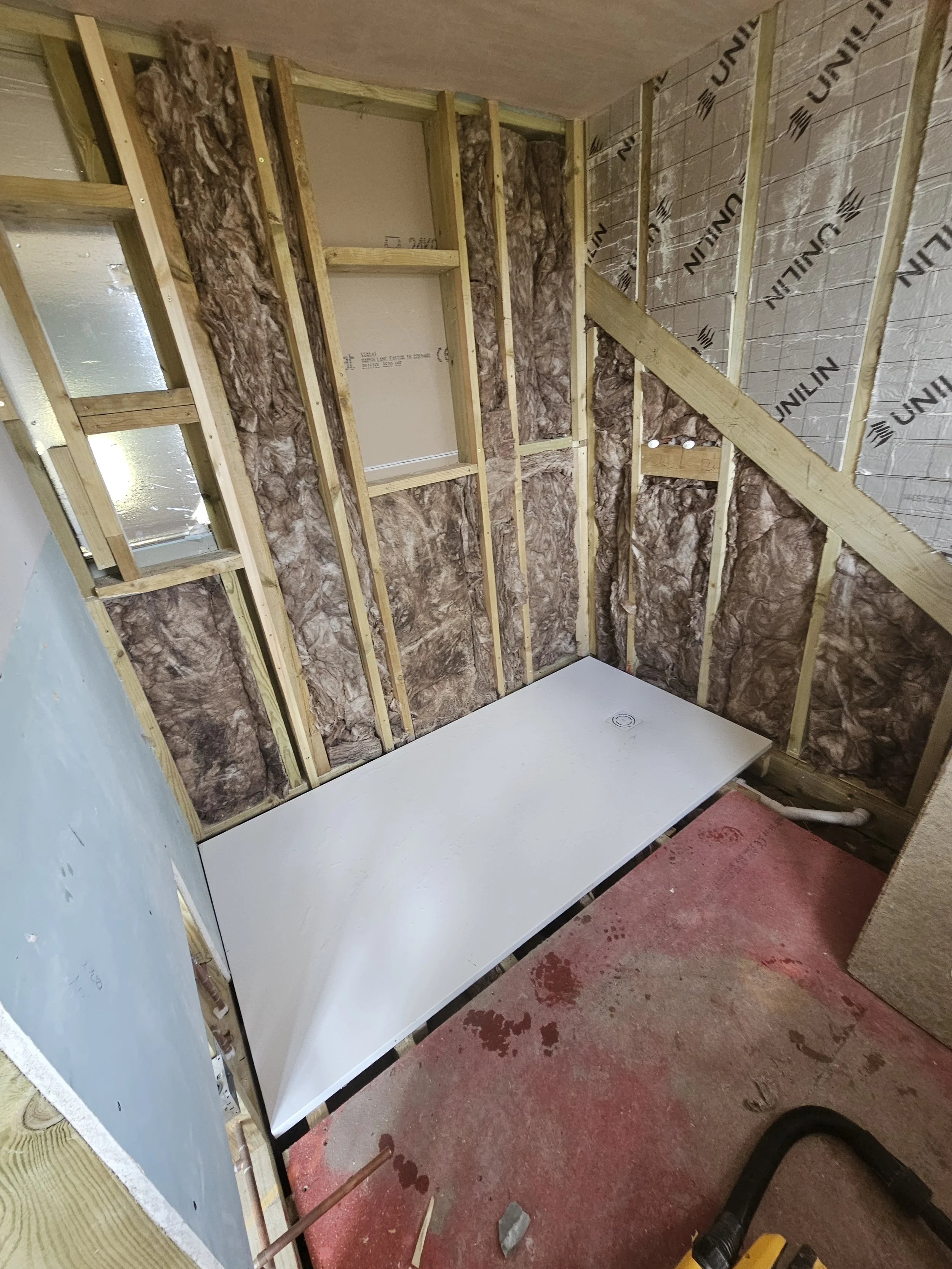 Unfinished room under construction showing exposed wooden framing, insulation, and a white panel on a red floor. The scene conveys a sense of progress.