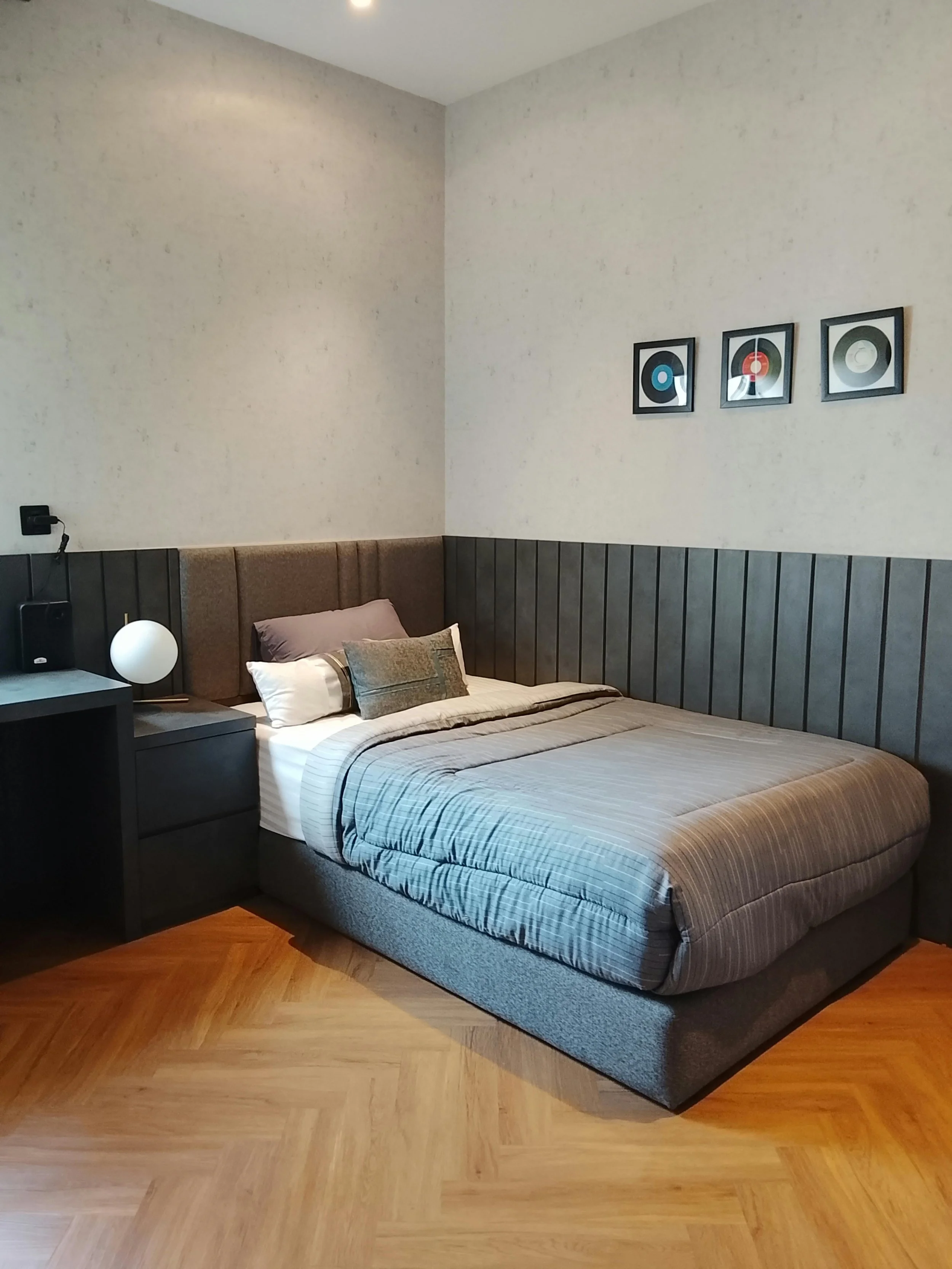 Cozy bedroom with a gray upholstered bed and nightstand. Soft lighting creates a calm atmosphere. Three framed records adorn the wall above.