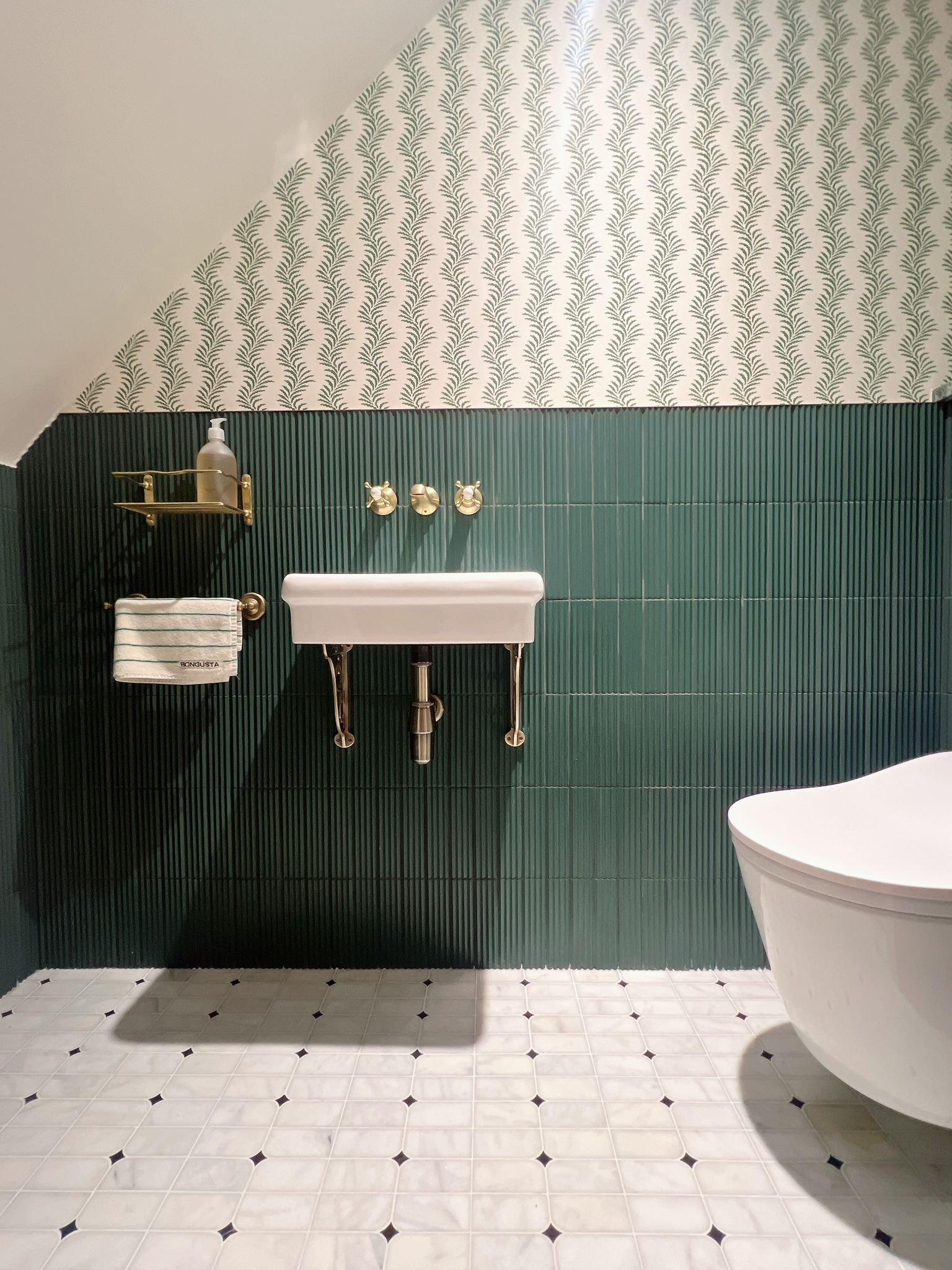 Bathroom with dark green vertical tiles, brass fixtures, a white sink, patterned wallpaper, and a round toilet. Clean and modern design.