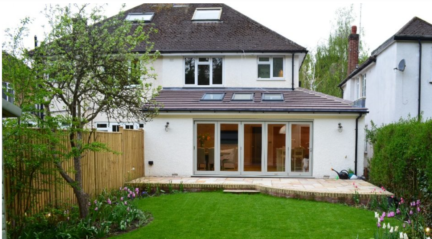 Two-story white house with a small patio. Glass doors lead to a bright interior. Bushes line the right side, and a chimney is visible. Tranquil setting.