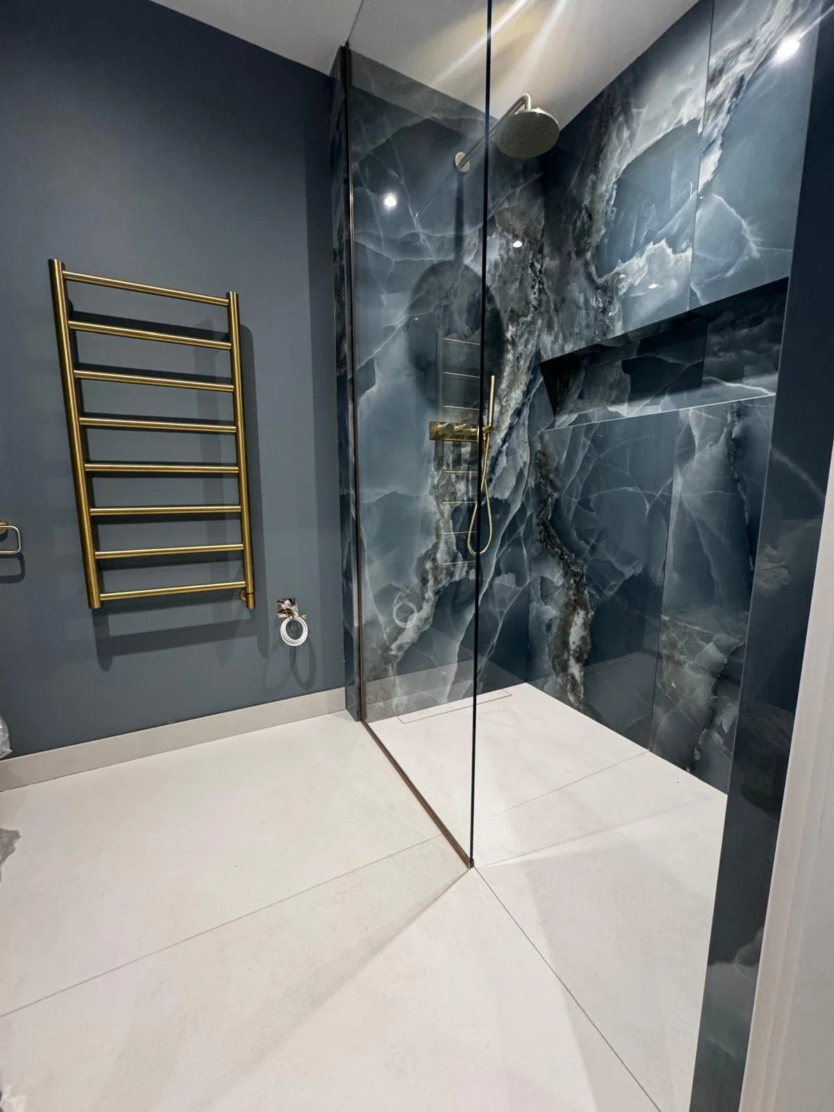 Modern bathroom featuring a glass-enclosed shower with dark marble-patterned walls, adjacent gold towel rack, and sleek light tiles. Stylish and elegant.