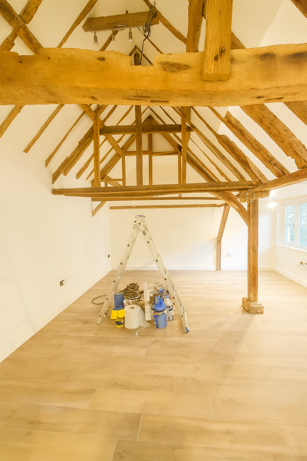 A bright, rustic room with exposed wooden beams and light wood flooring. A stepladder stands in the center, surrounded by paint supplies.