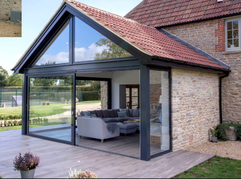 Modern house with large glass doors and a triangular window, opening to a green garden and tennis court. Indoor area features a cozy grey sofa, conveying tranquility.