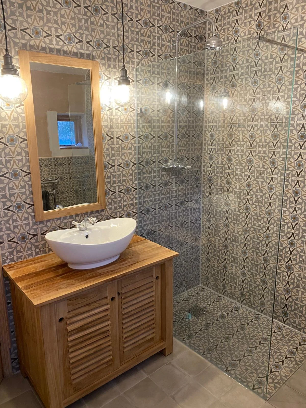 A cozy bathroom features a wooden-framed mirror, a round white sink on a wooden vanity, patterned tiles, a glass shower screen, and hanging light bulbs.