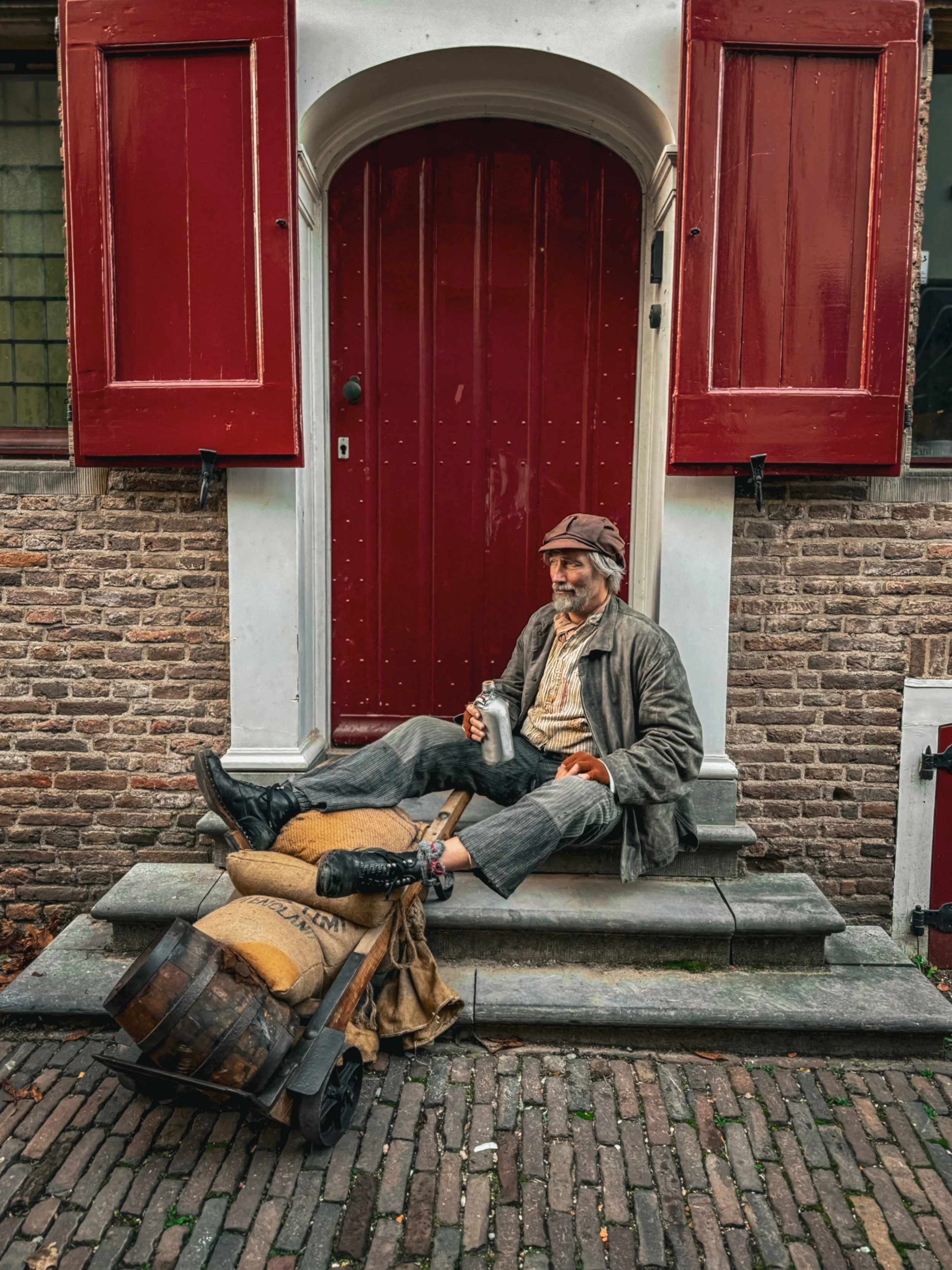 Older man with gray hair and beard, sitting on steps outside a house with red door and shutters, holding a metal mug, surrounded by bags and a hand truck.