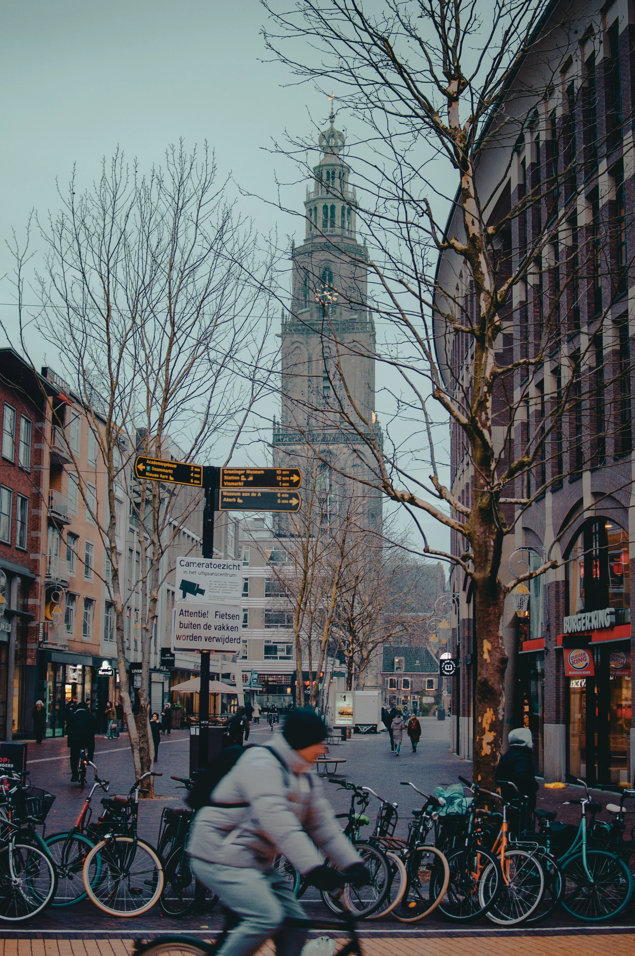 A city street scene with leafless trees, bicycles, pedestrians, and a tall historic church tower in the background.