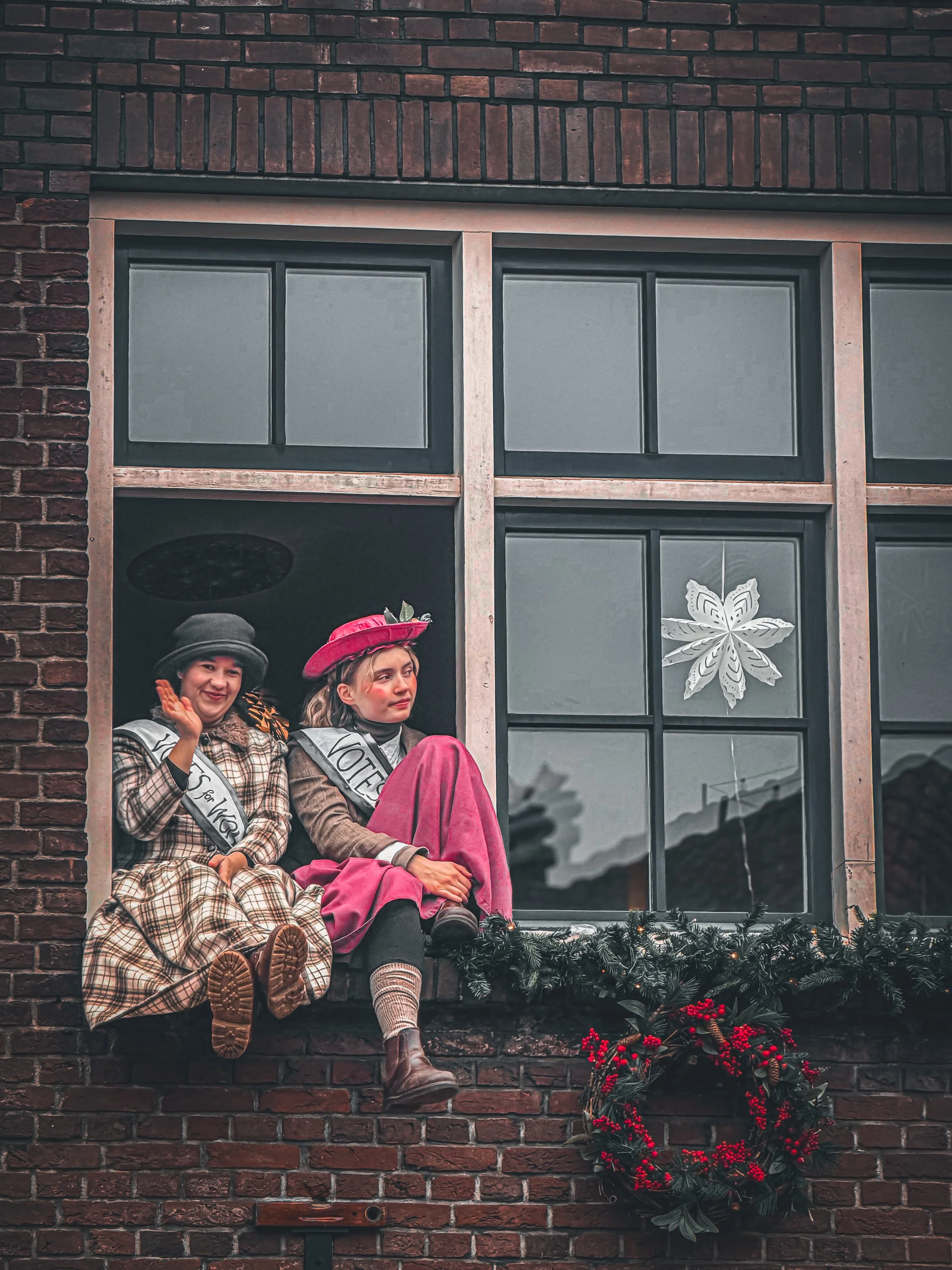 Two women dressed as historical figures sitting on a window ledge decorated with holiday garlands and wreaths, with a snowflake ornament on the window, during a festive event.