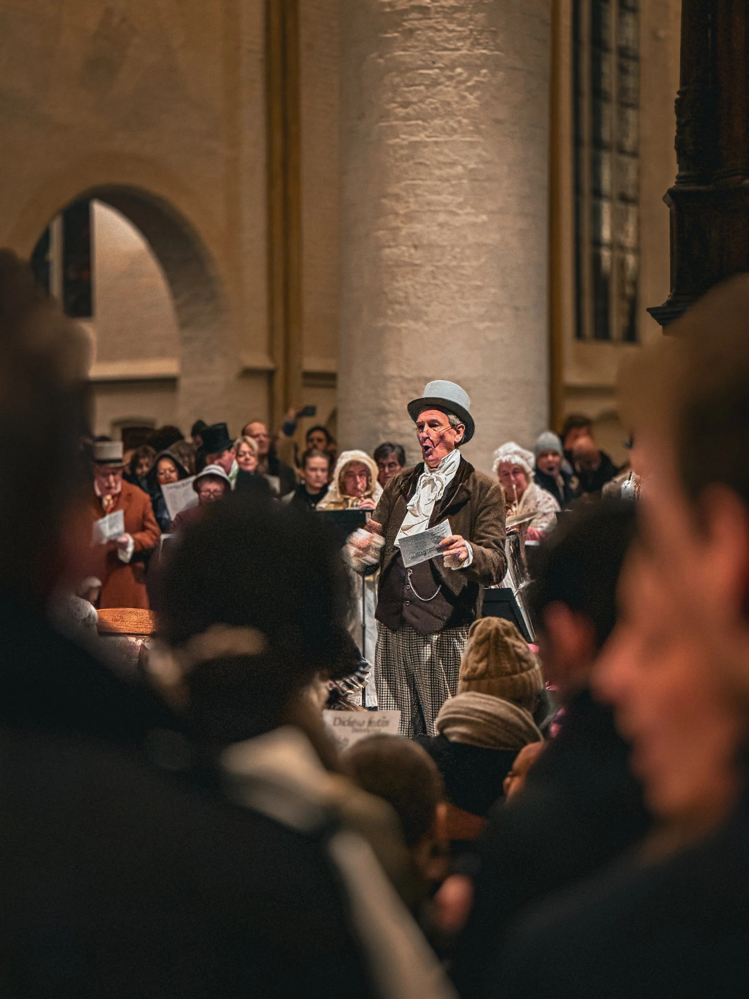 A group of people dressed in historical clothing are gathered in a church or similar building, with one woman in the center dressed in 19th-century attire singing or speaking to the crowd.