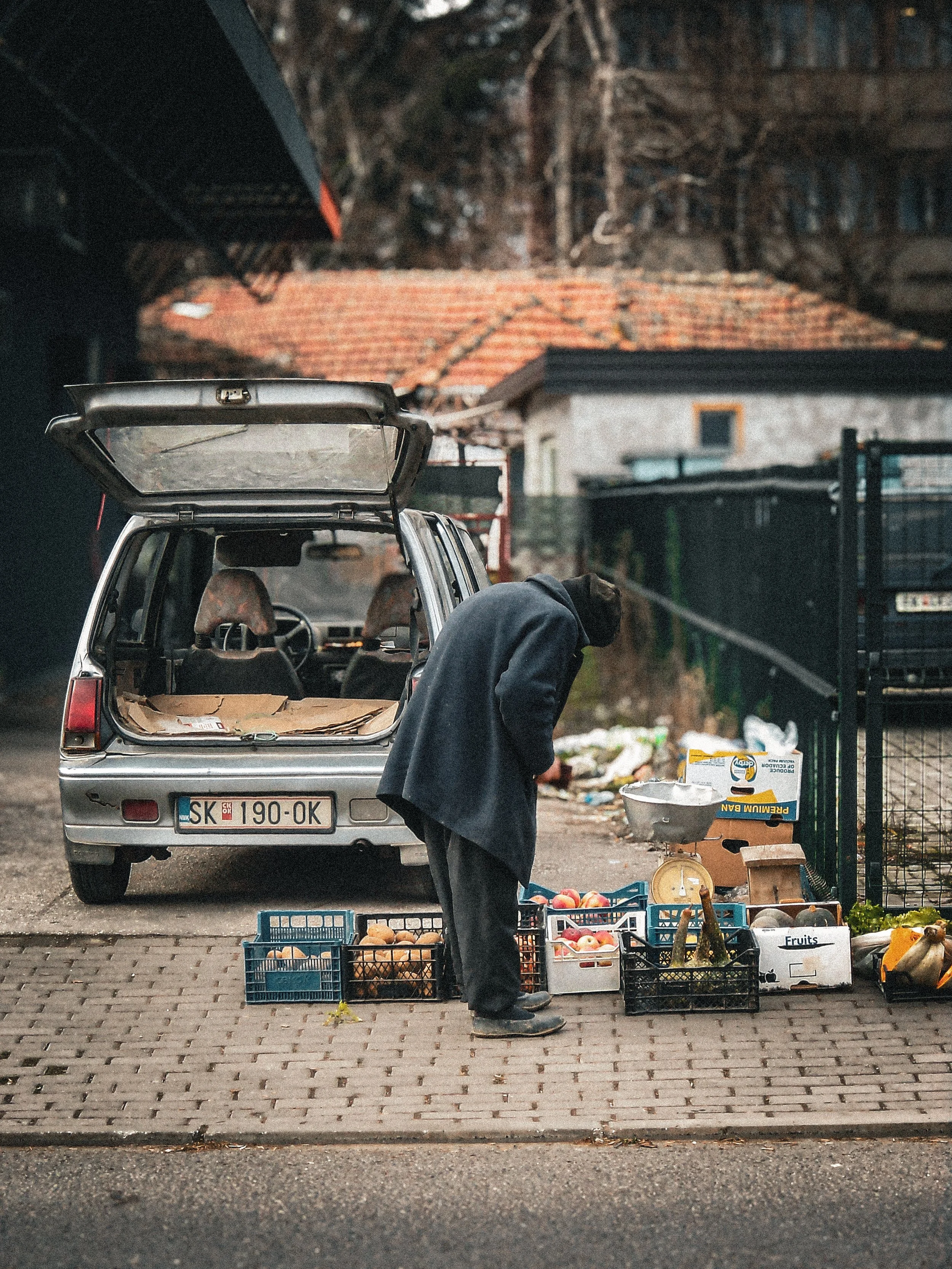 A man at a street market stall with crates of fruits and vegetables, standing in front of an open car trunk, on a sidewalk in an urban area with houses and trees in the background.
