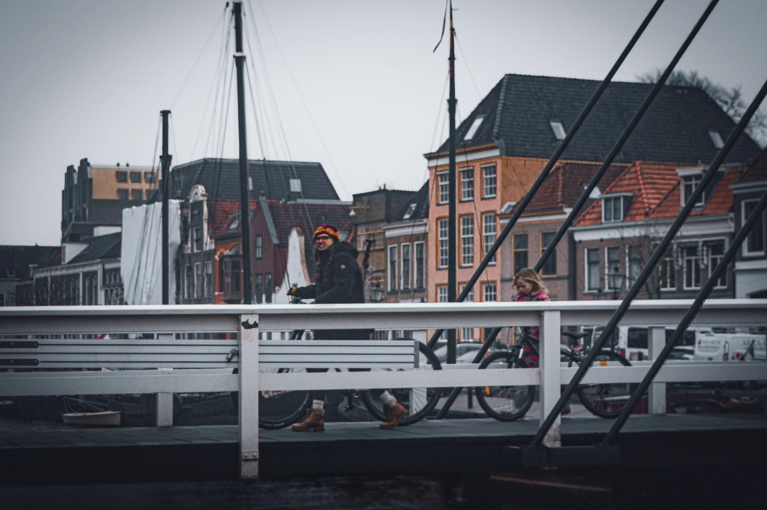 People walking their bicycles on a bridge in a town with colorful buildings in the background.