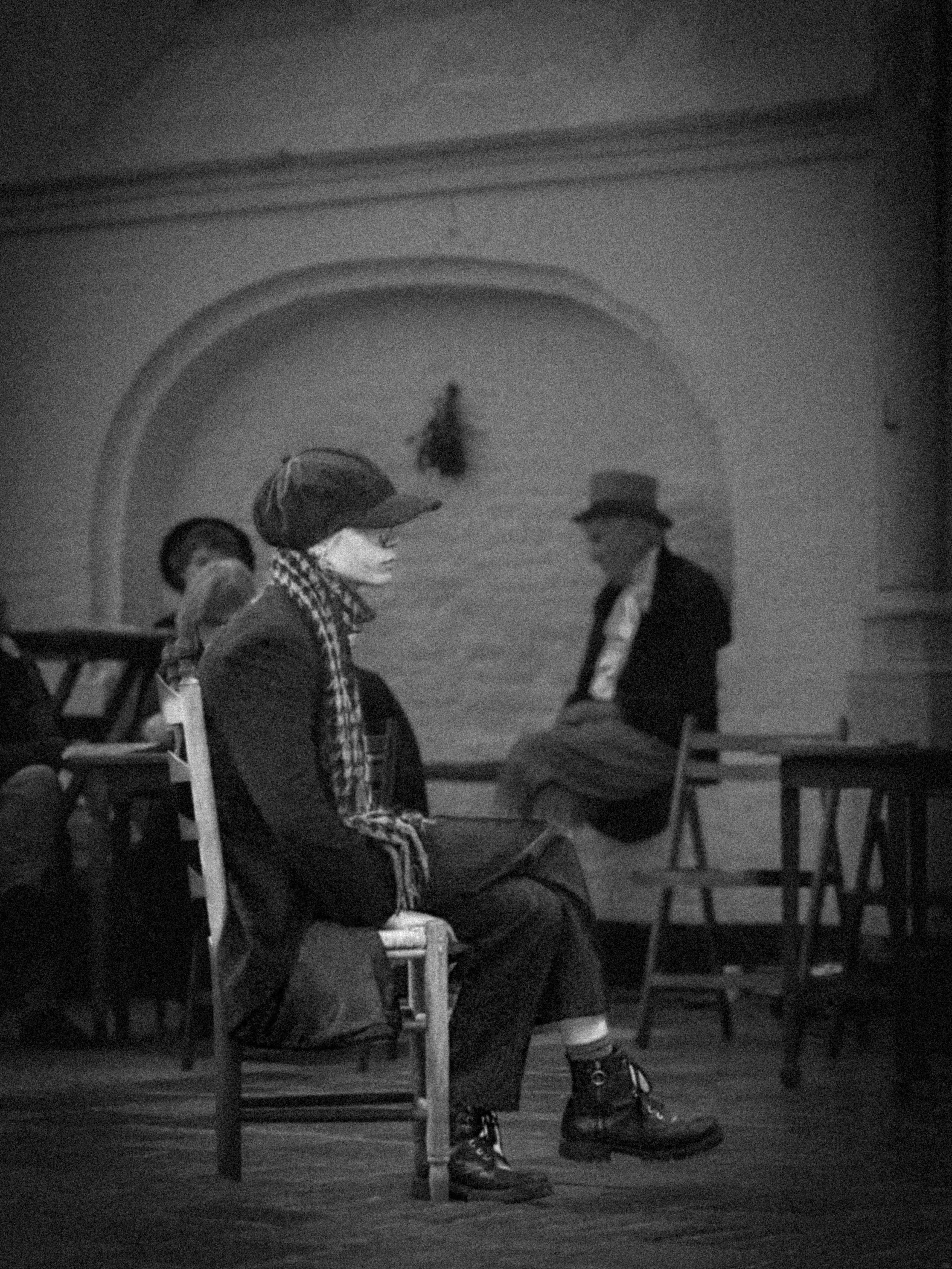 People sitting in a room, some wearing hats and jackets, with a large arched opening in the background.