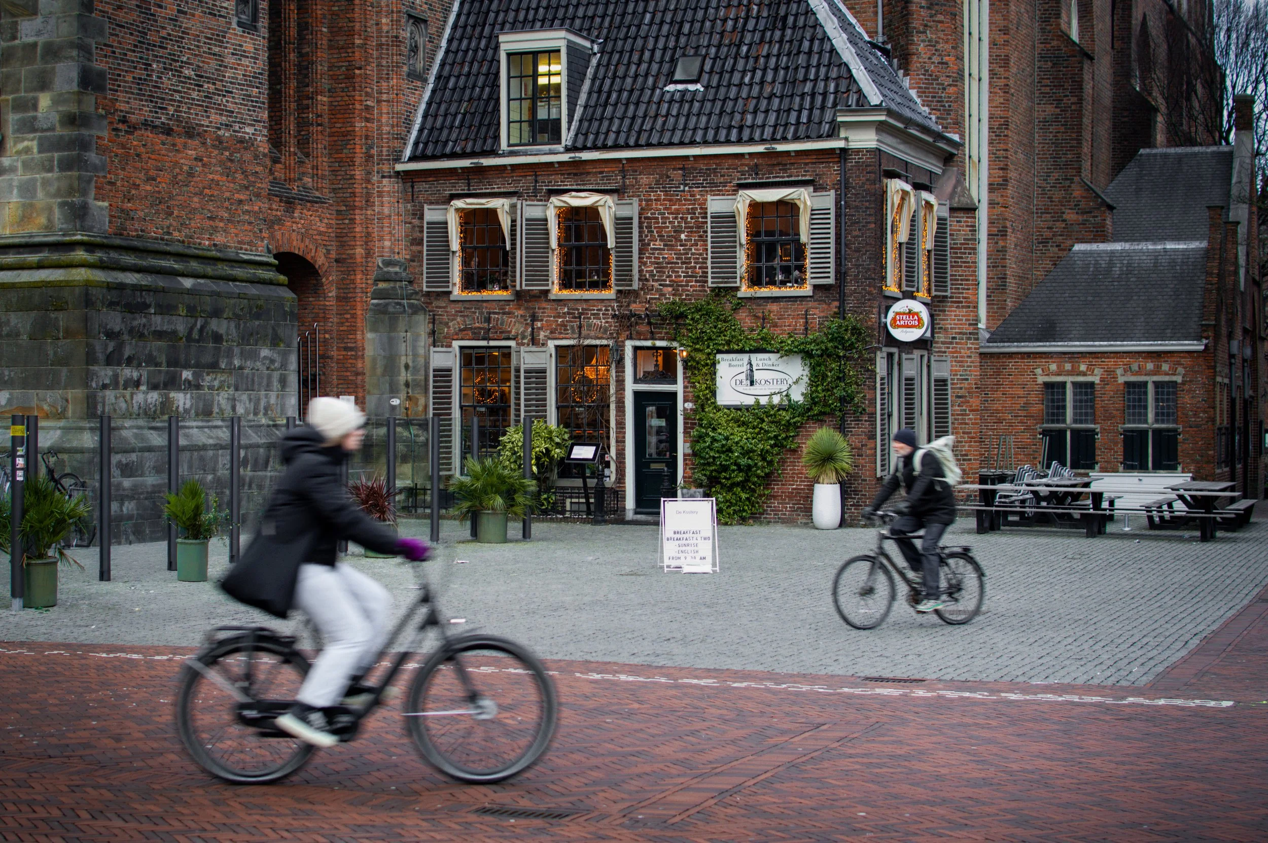 Two people riding bicycles past a brick building with a sign that reads 'De Kustery'. The building has multiple windows with white shutters and a sign on the wall for Stella Artois. There are potted plants and outdoor seating on a patio area.