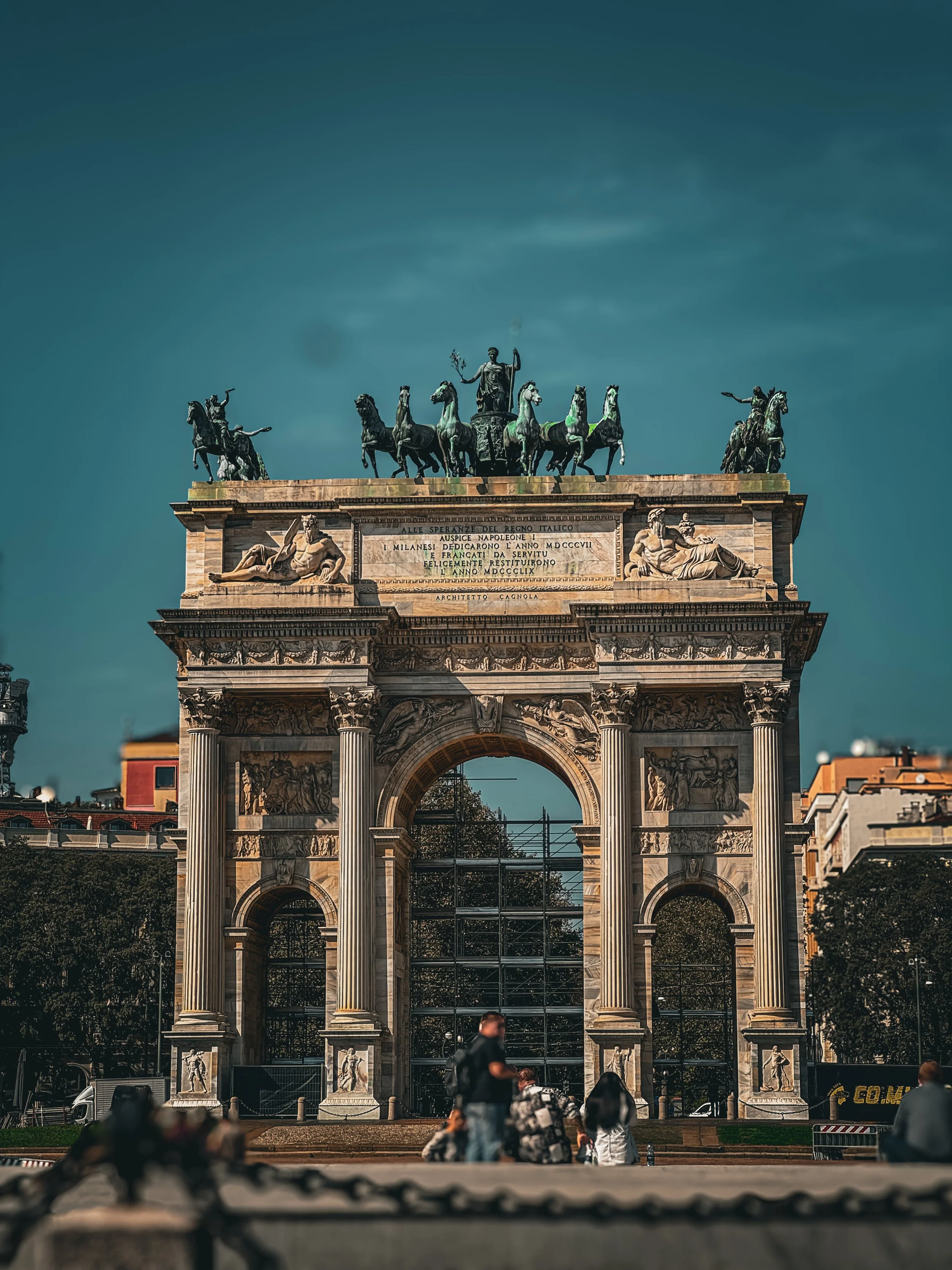 The Arco della Pace, a large arch with sculptures and a chariot statue on top, is a historic monument in Milan, Italy. People are seated and walking in front of it.