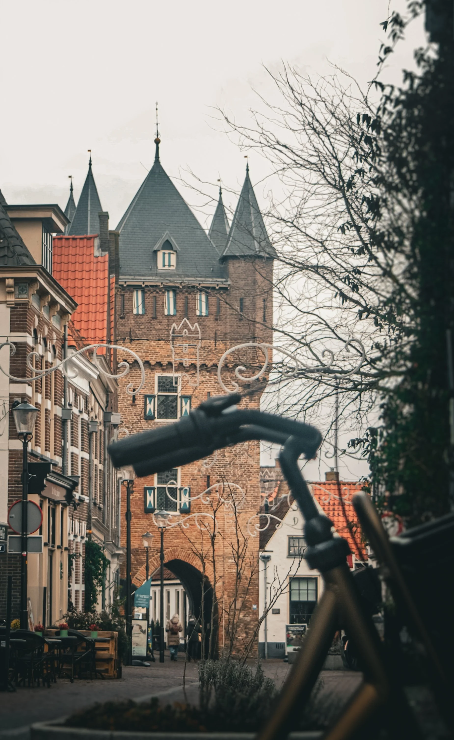 A historic brick tower with pointed gray roofs and narrow arched windows in a European city street, with leafless trees and a bicycle in the foreground.