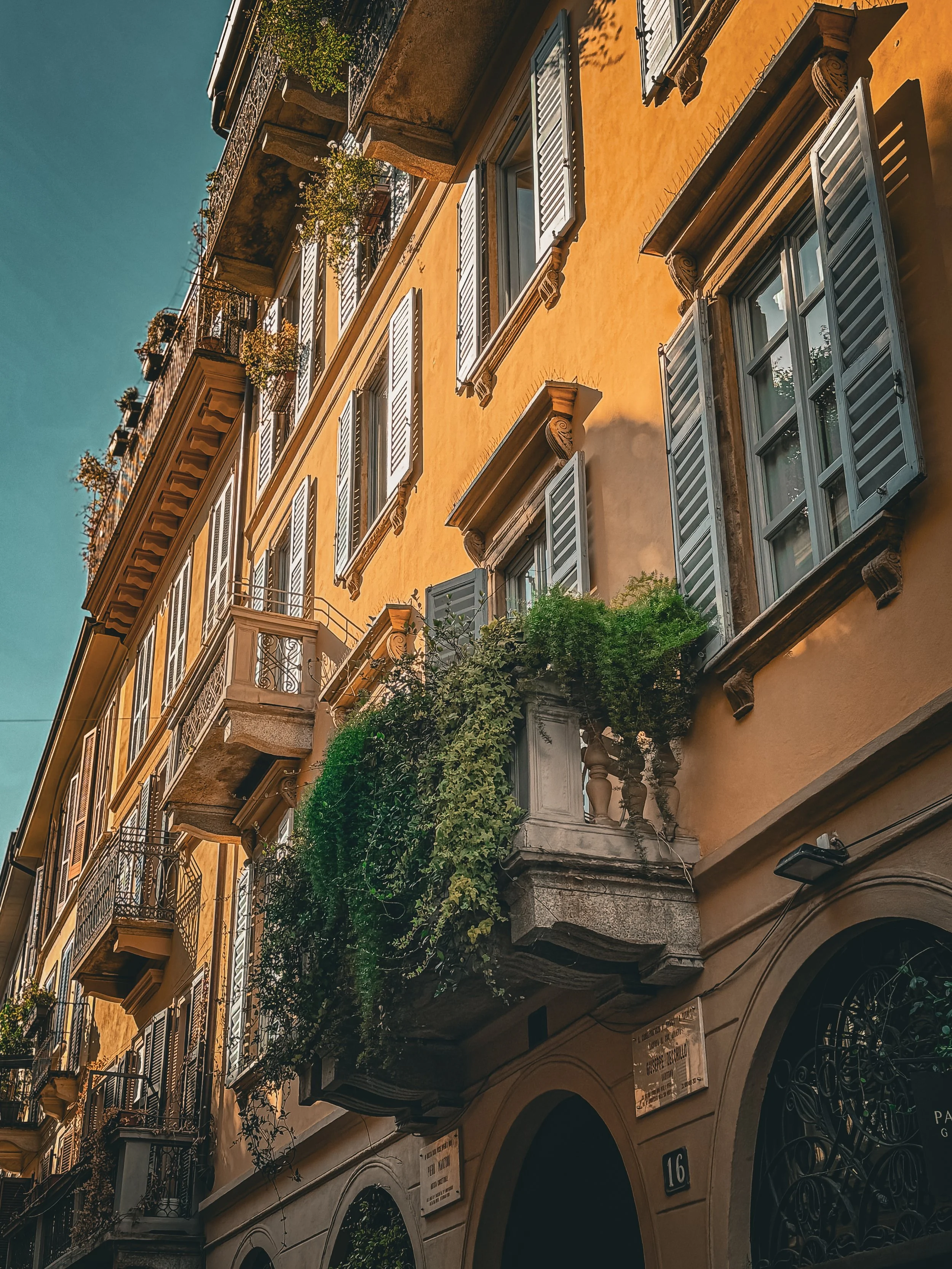 A multi-story yellow building with open white window shutters, balconies with greenery, and ornate architectural details in what appears to be a European city.