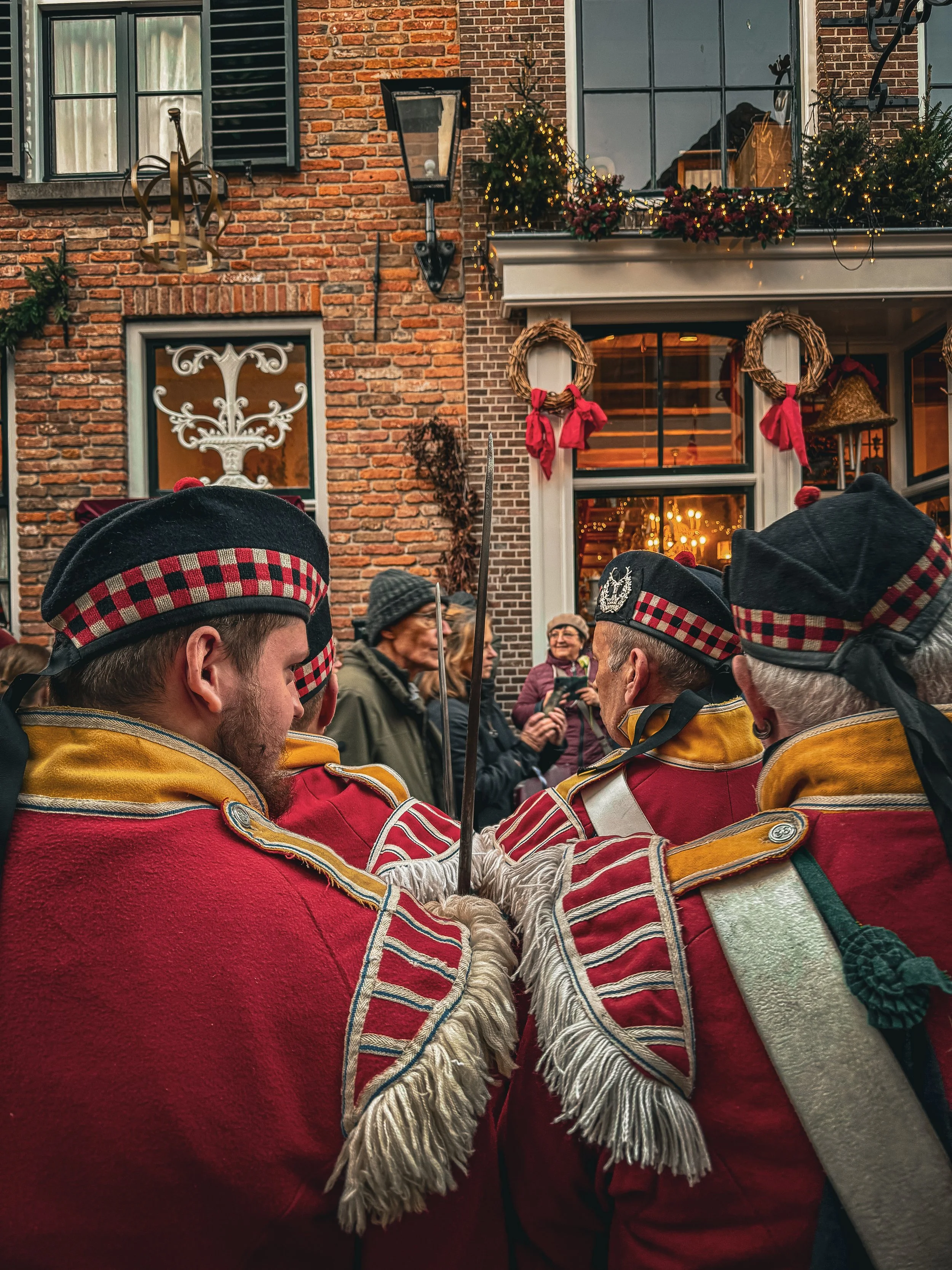 Historical reenactors dressed as soldiers in red uniforms and black hats with checked bands, engaging in a ceremonial activity during a Christmas celebration in front of a decorated brick building, with onlookers and festive holiday decorations.