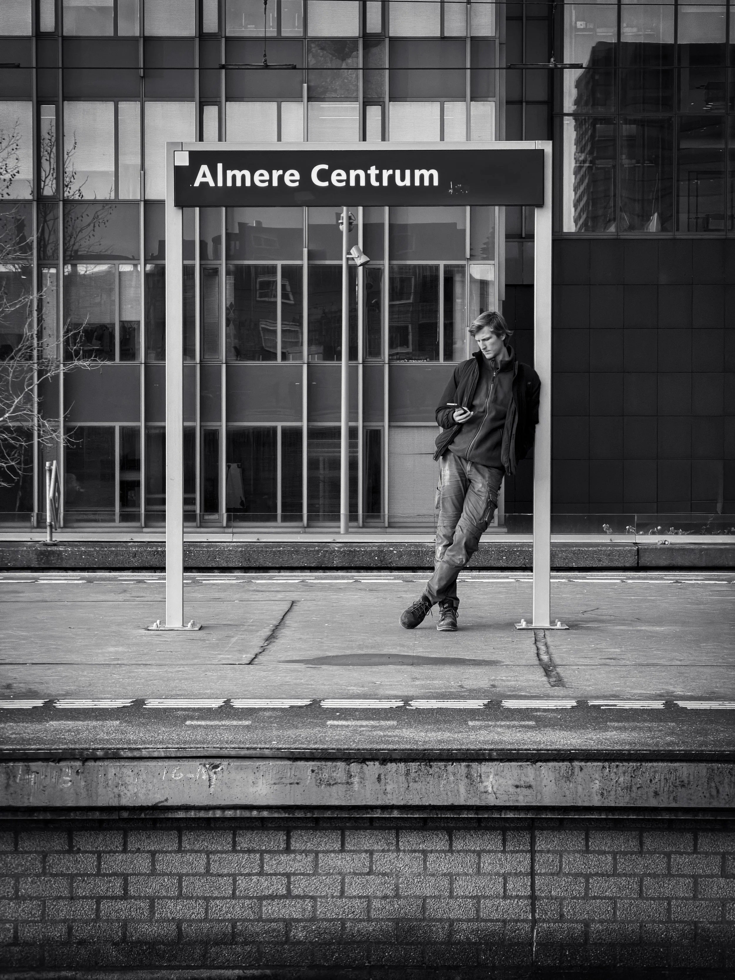 A young man leaning against a signpost at Almere Centrum train station. He is looking at his phone, dressed in casual clothing, with a modern glass building in the background.