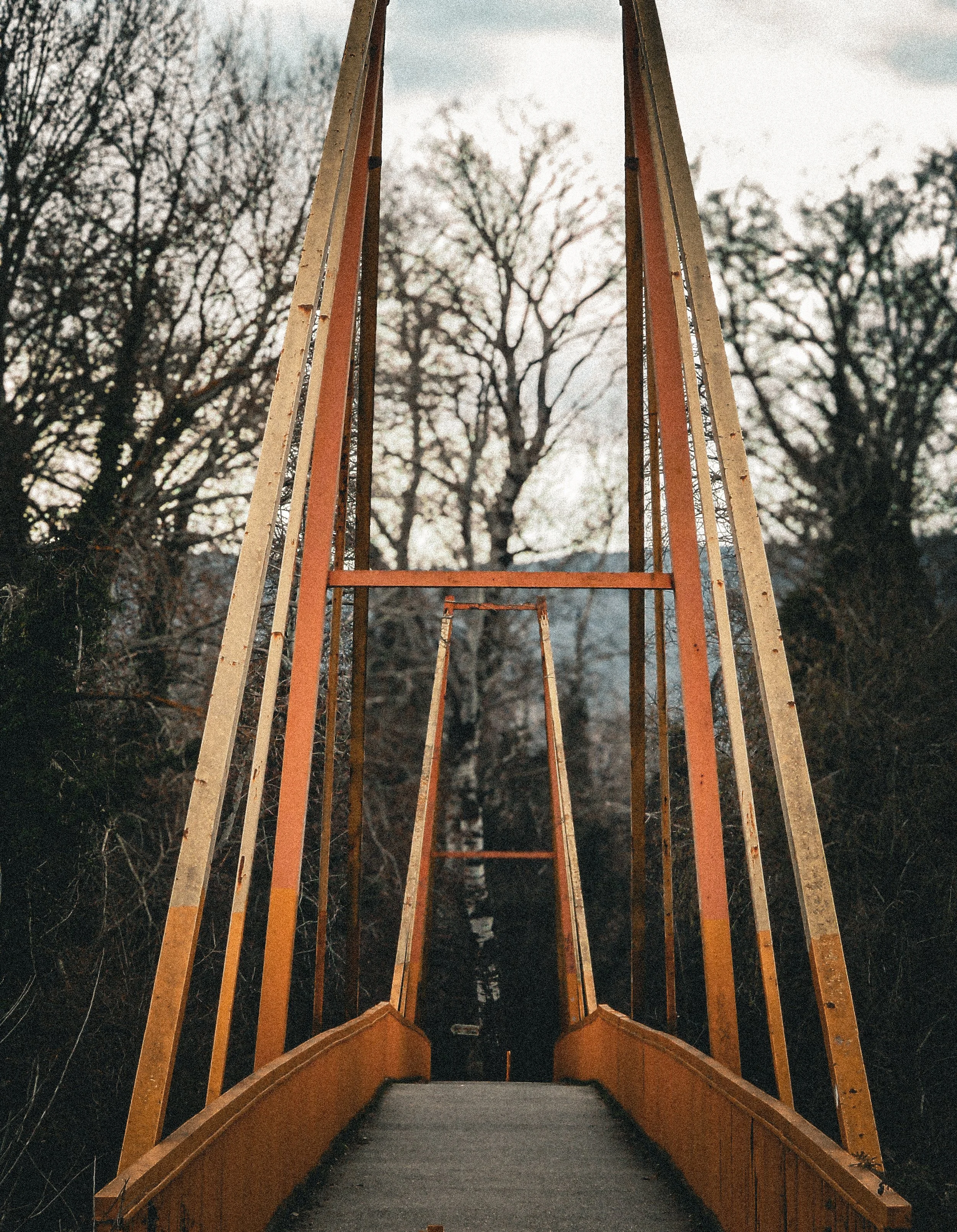 An empty yellow suspension bridge with orange support beams extending into the distance, surrounded by leafless trees and overcast sky.