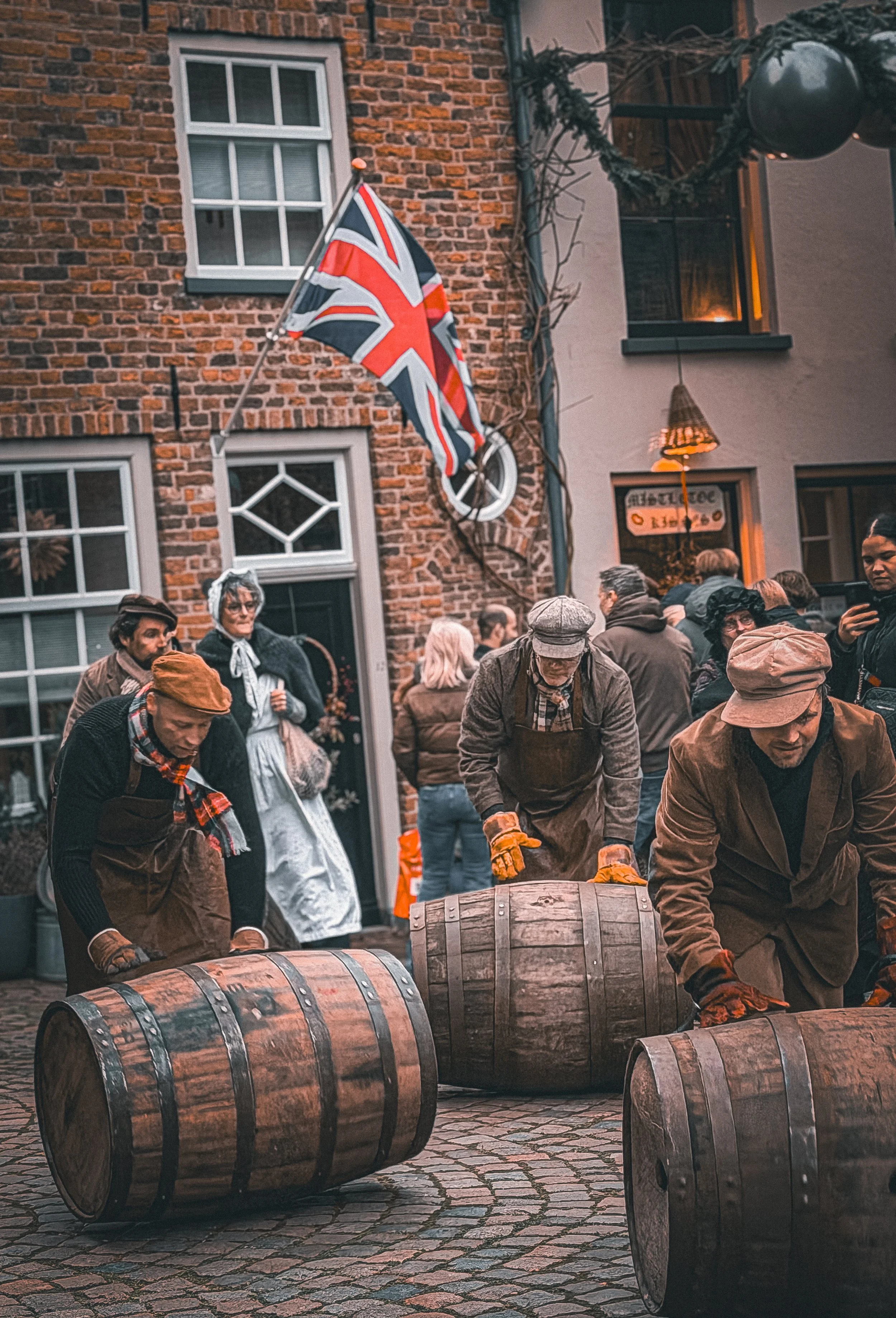 People dressed in vintage clothing rolling wooden barrels on cobblestone street during a historical reenactment.