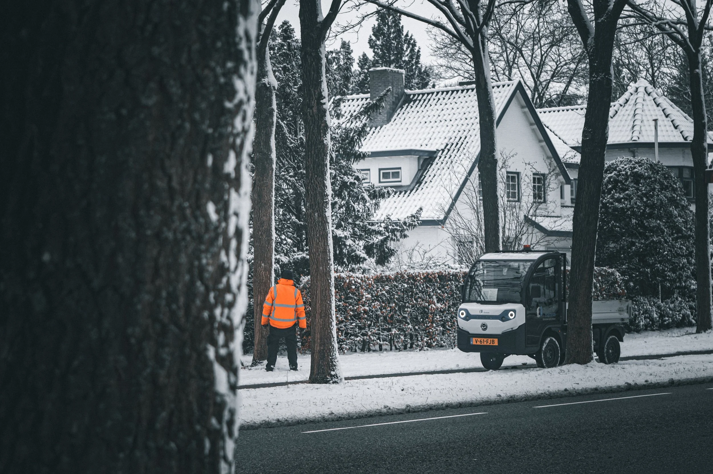 Snow-covered street with a person in an orange safety vest working near a small utility vehicle, surrounded by trees and houses with snow on their roofs.