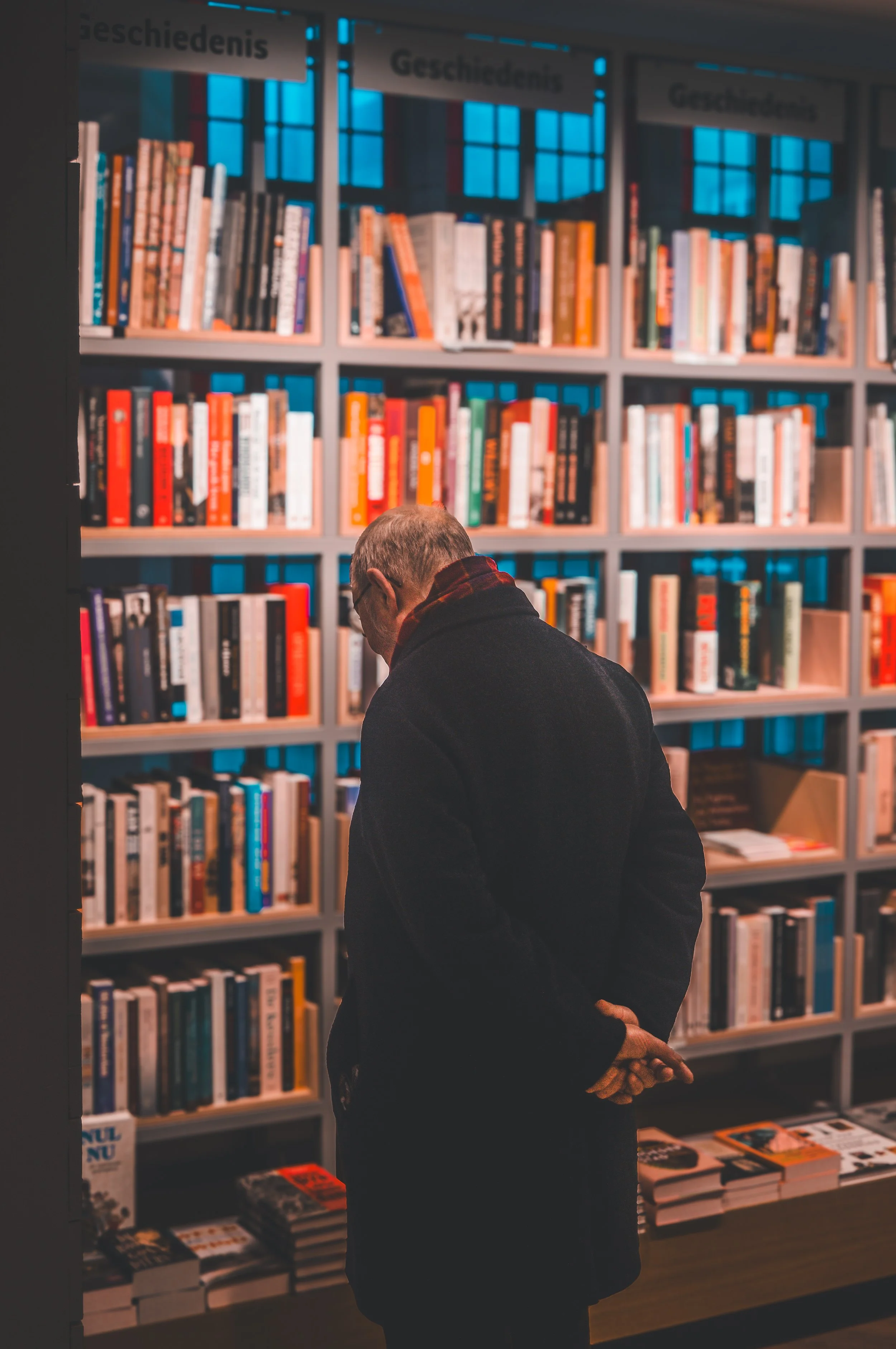 An older man stands in front of a bookshelf filled with colorful books in a library or bookstore, with his hands clasped behind his back and head slightly bowed, reading or contemplating.