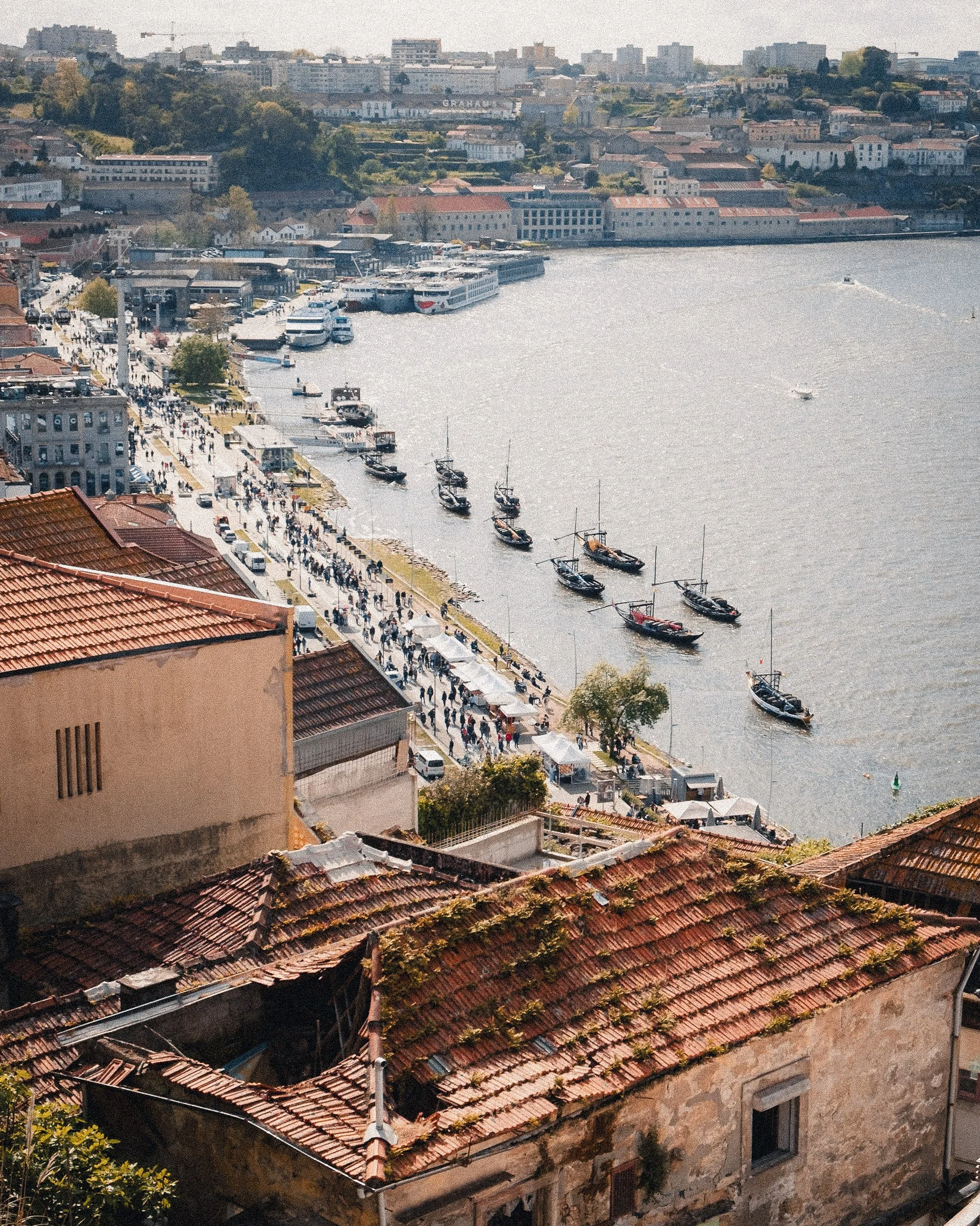 Aerial view of a riverside promenade with boats docked along the shore, crowded with pedestrians, lined by old and modern buildings, with a hillside in the background.