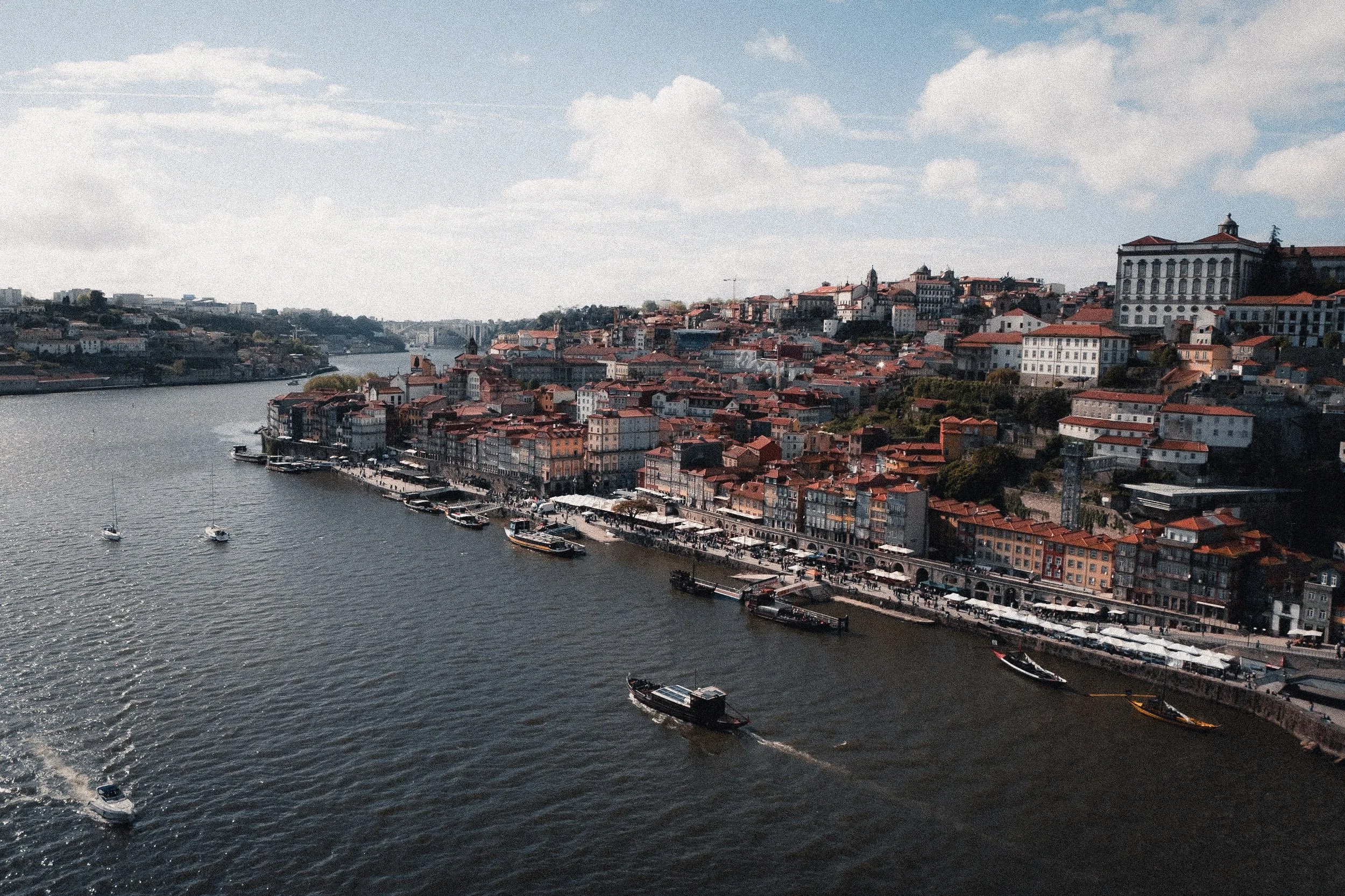 Aerial view of a riverside city with boats on the water, colorful buildings lining the shore, and a hill with houses and trees in the background, under a partly cloudy sky.