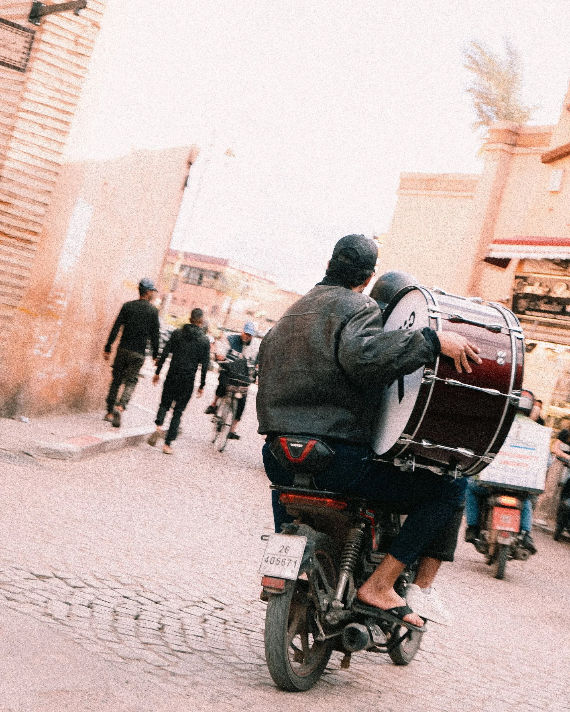 Man riding a motorcycle with a bass drum attached, moving through a city street with pedestrians and cyclist.