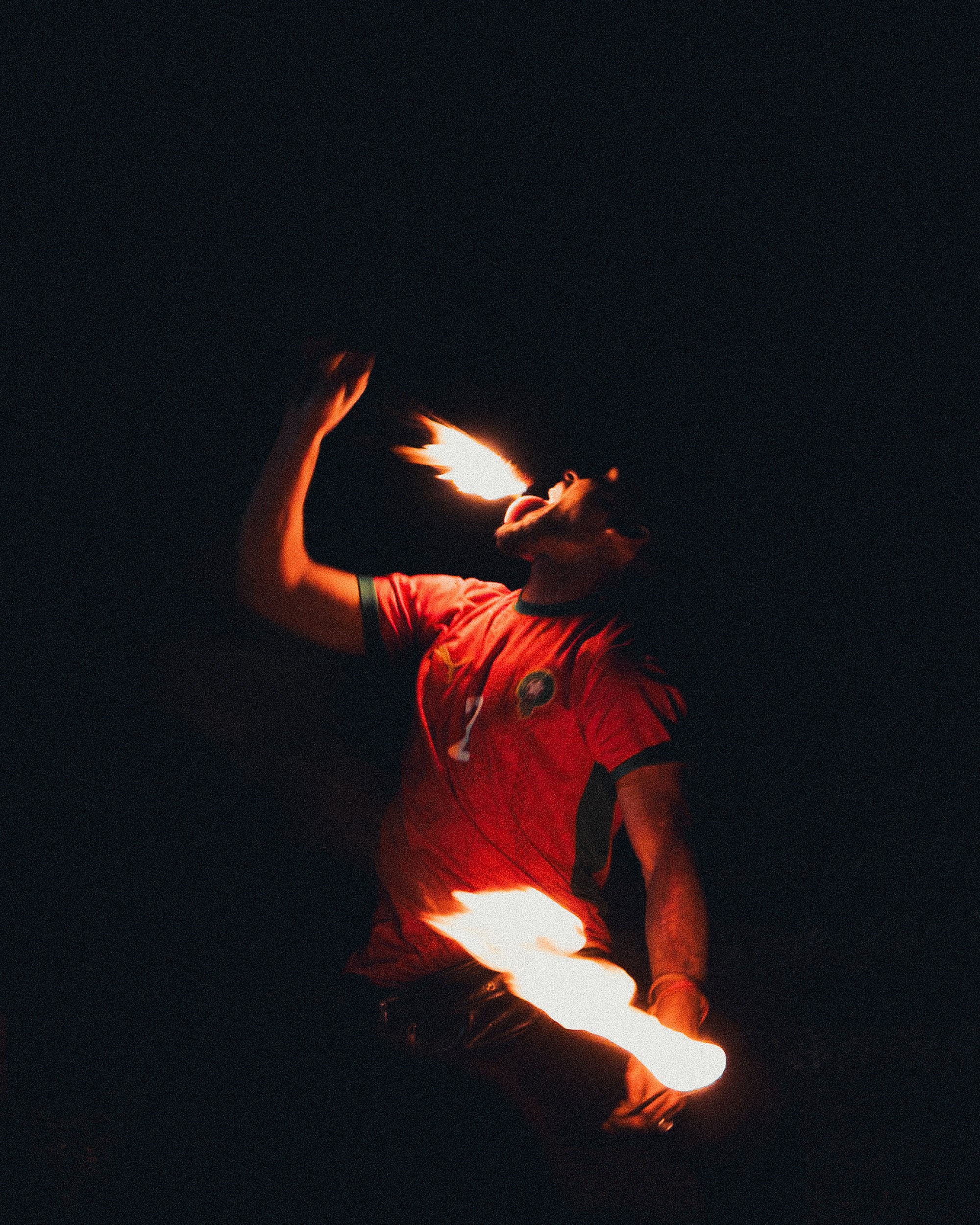 Person performing fire breath trick at night, wearing a red sports jersey with a crest, holding a flaming object in one hand and breathing fire.