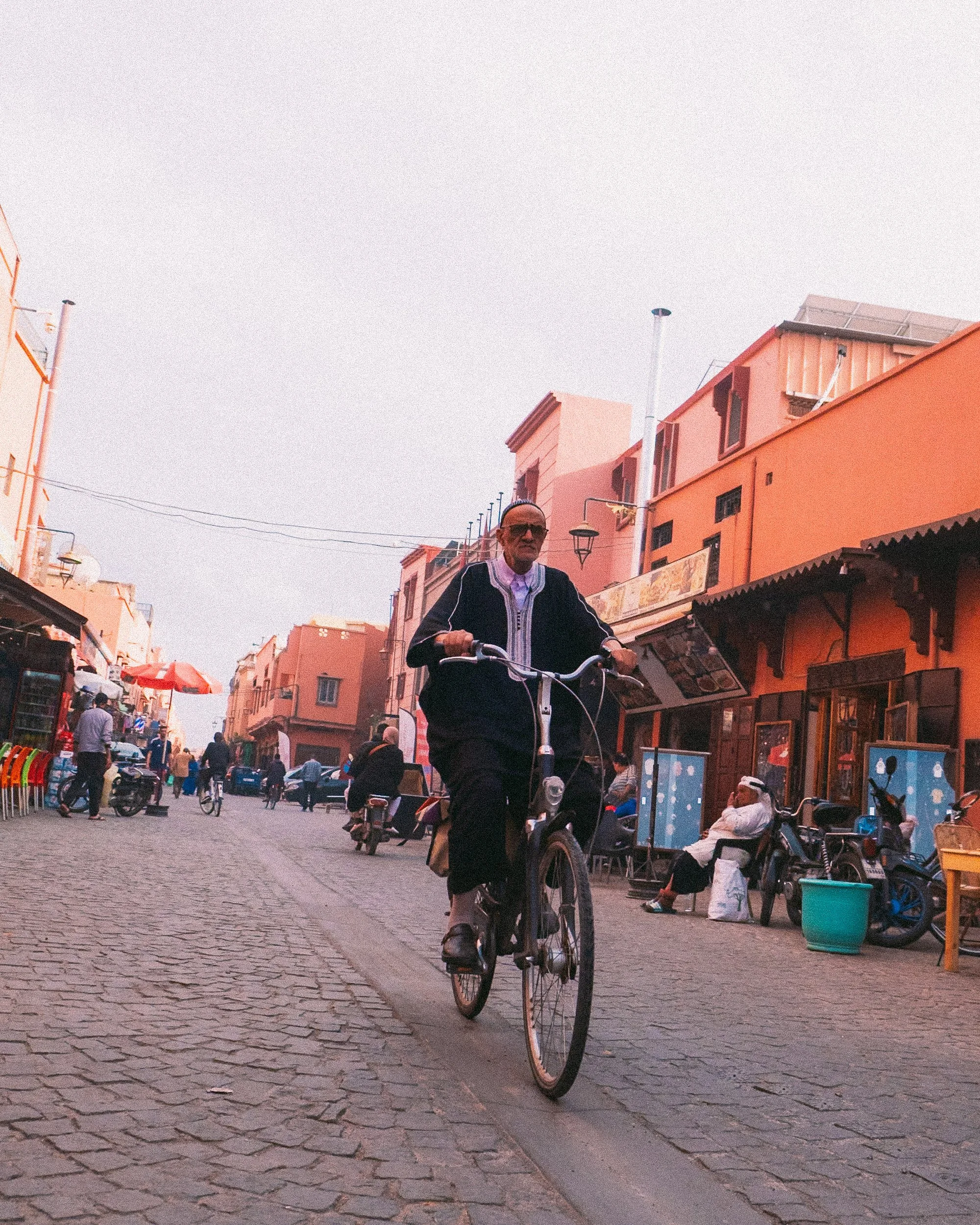 A man riding a bicycle down a cobblestone street in a lively market area with shops and pedestrians.