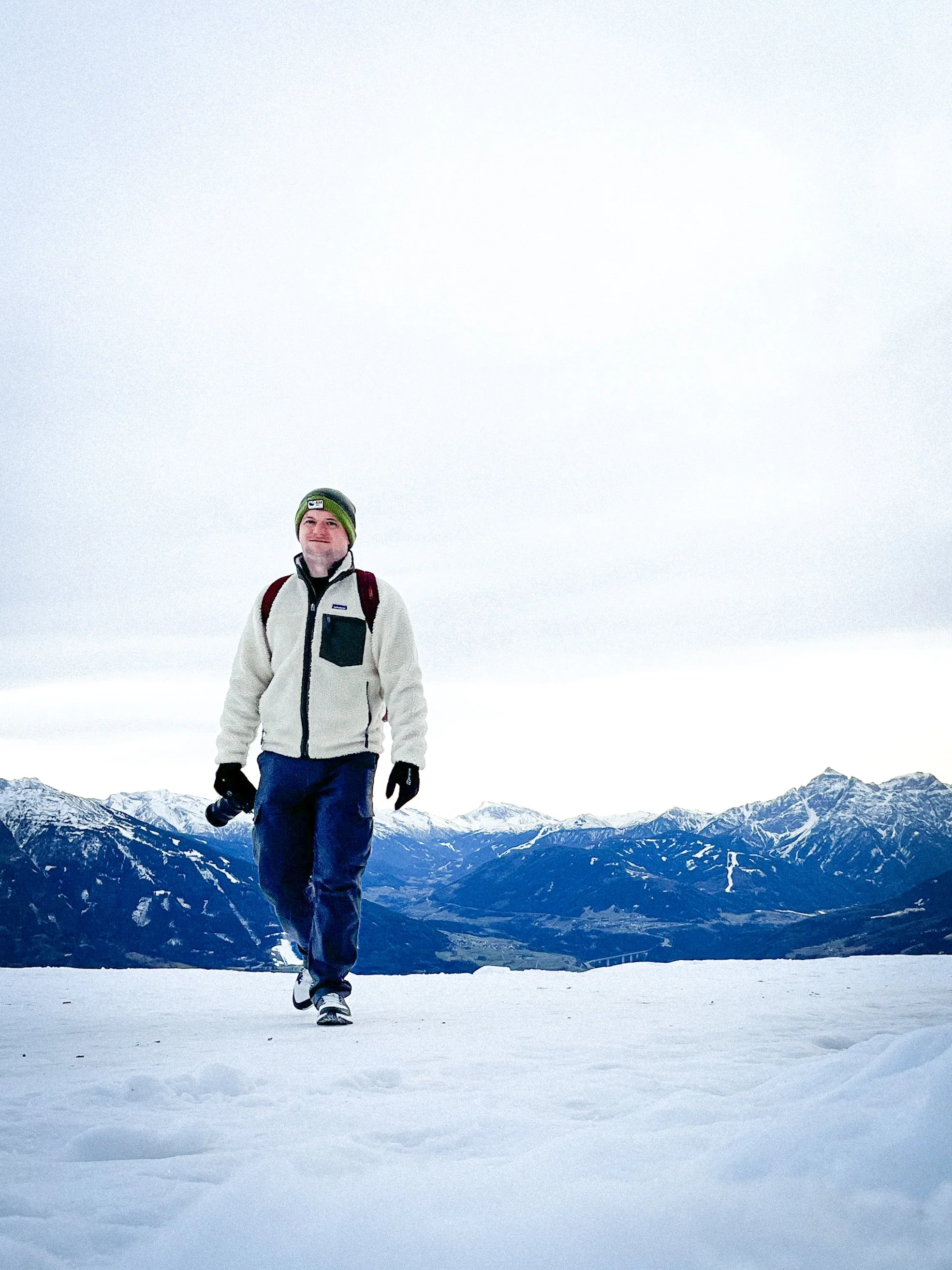 A man walking on snow-covered ground in a mountainous landscape, wearing a white jacket, blue pants, a green beanie, and carrying a backpack, with snowy mountains in the background.