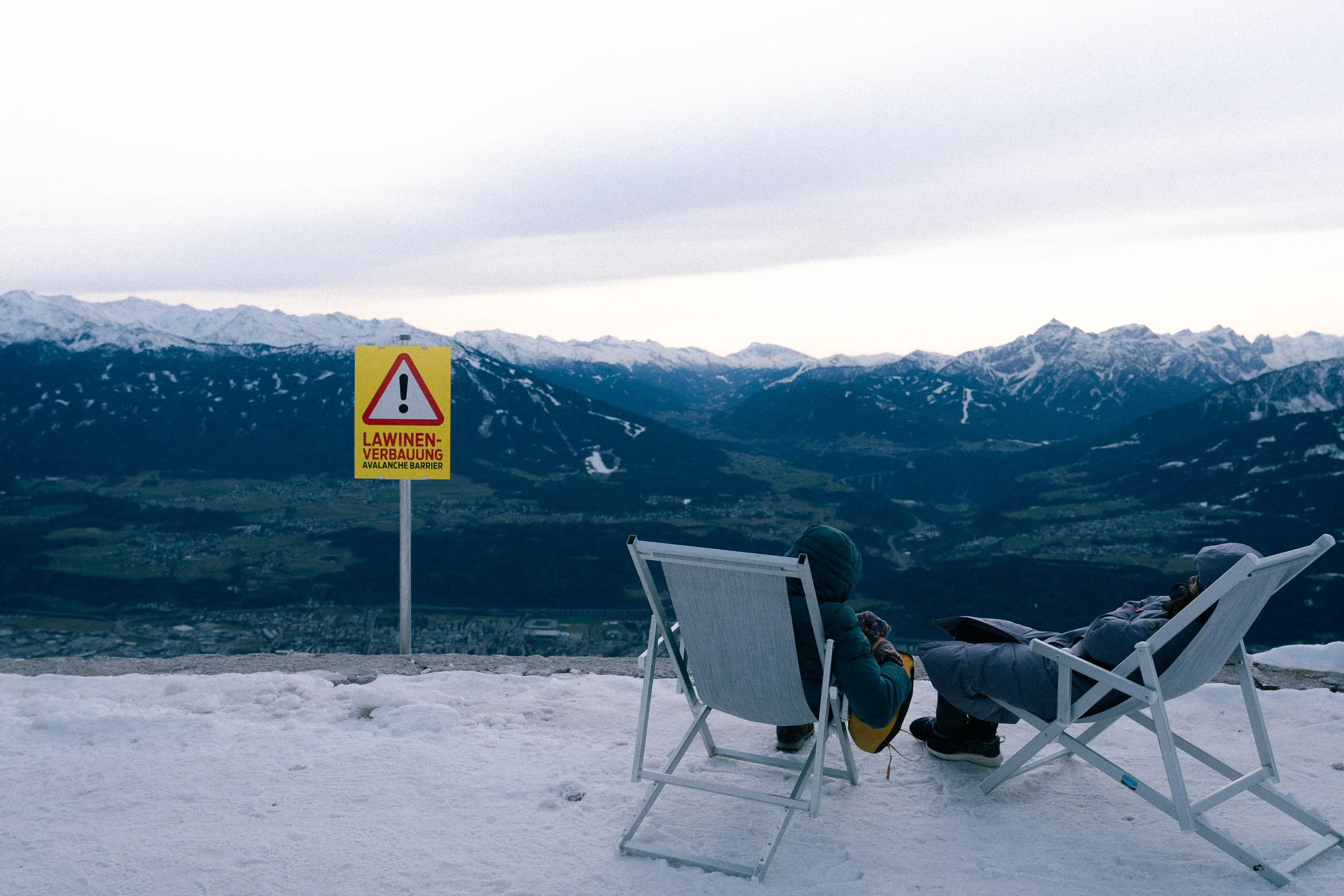 Two people are sitting in chairs on a snowy mountainside, facing a mountain range with snow-capped peaks, a sign warning about an avalanche barrier, overcast sky.