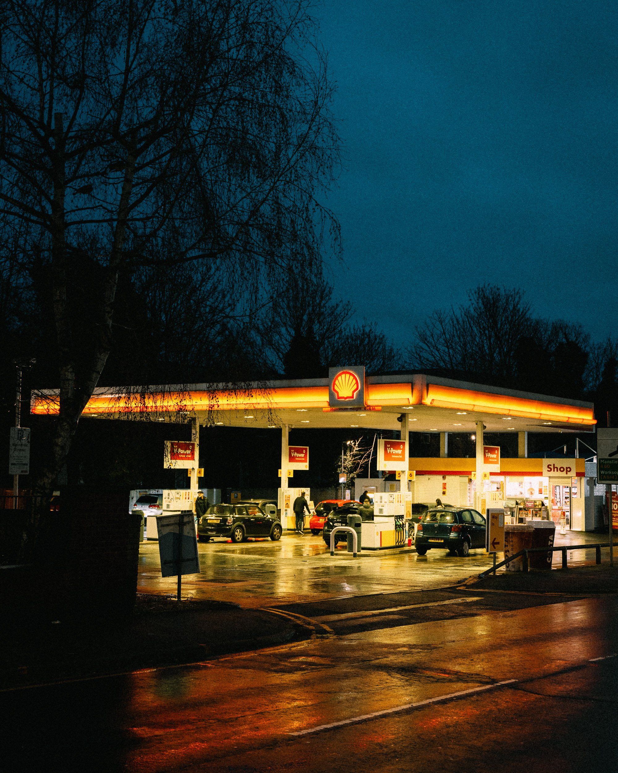 A Shell gas station illuminated at night with cars parked at the pumps and a shop in the background.