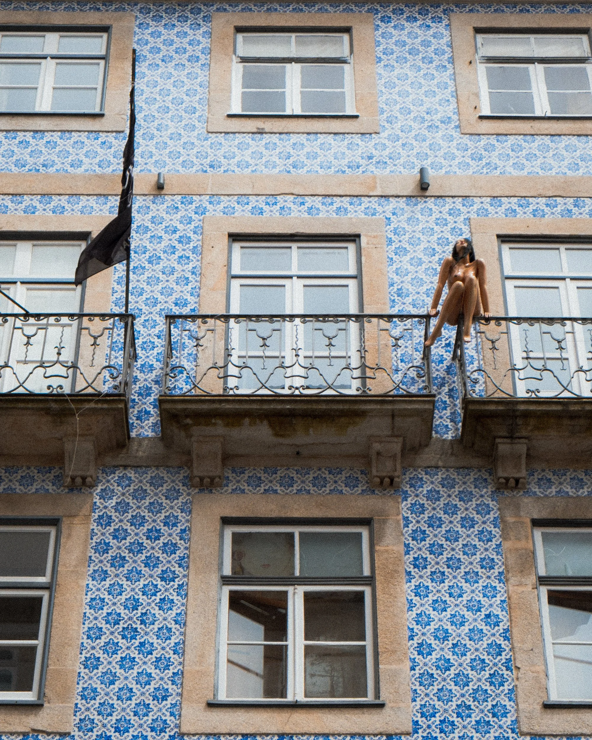 A woman sitting on a balcony of a building with blue and white patterned tile facade, looking up with sunglasses and long hair.