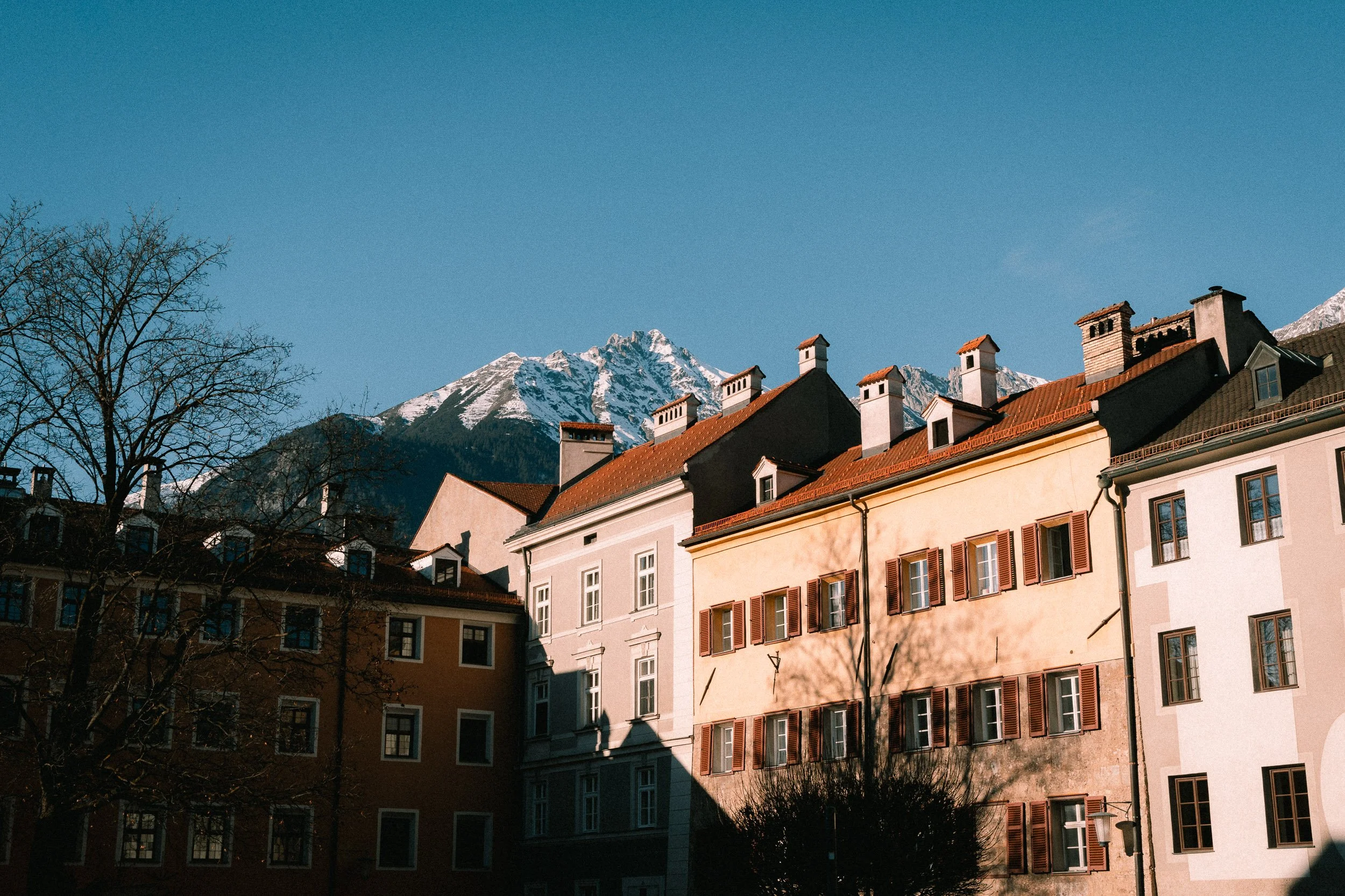 Colorful European-style houses with red-tiled roofs in front of snow-capped mountains under a clear blue sky.