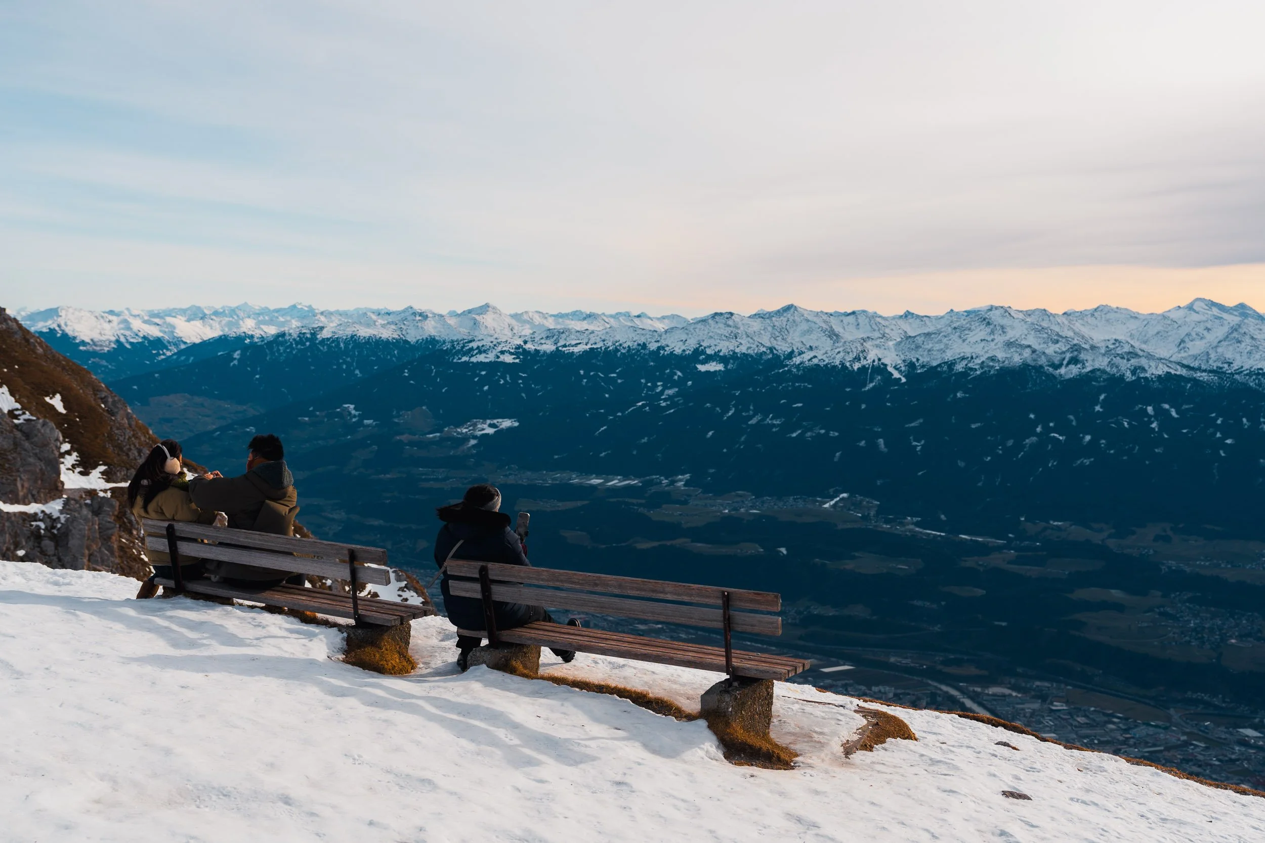Four people sitting on benches on a snow-covered mountain overlook, with snow-capped mountains and a cloudy sky in the background.