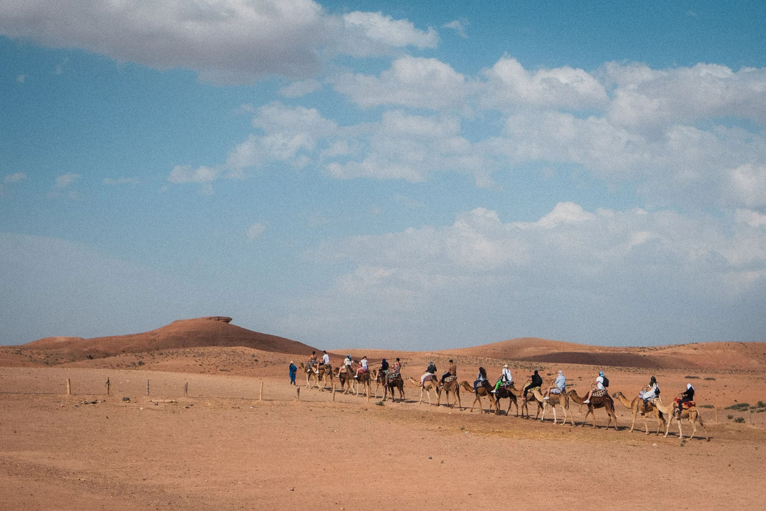 A group of tourists riding camels in a desert landscape under a partly cloudy sky.