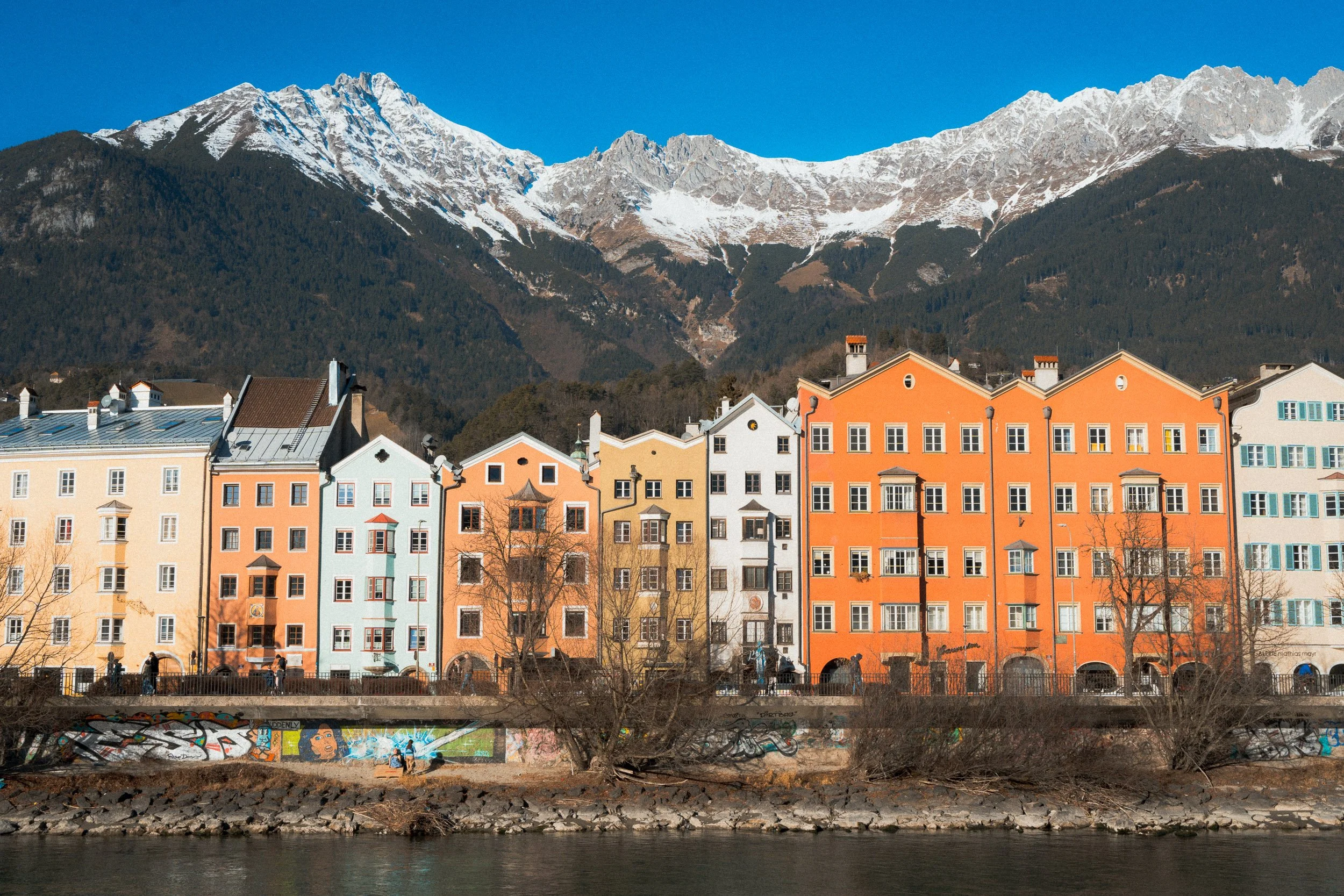 Colorful alpine town with pastel buildings along a river, snowy mountains in the background, and clear blue sky.