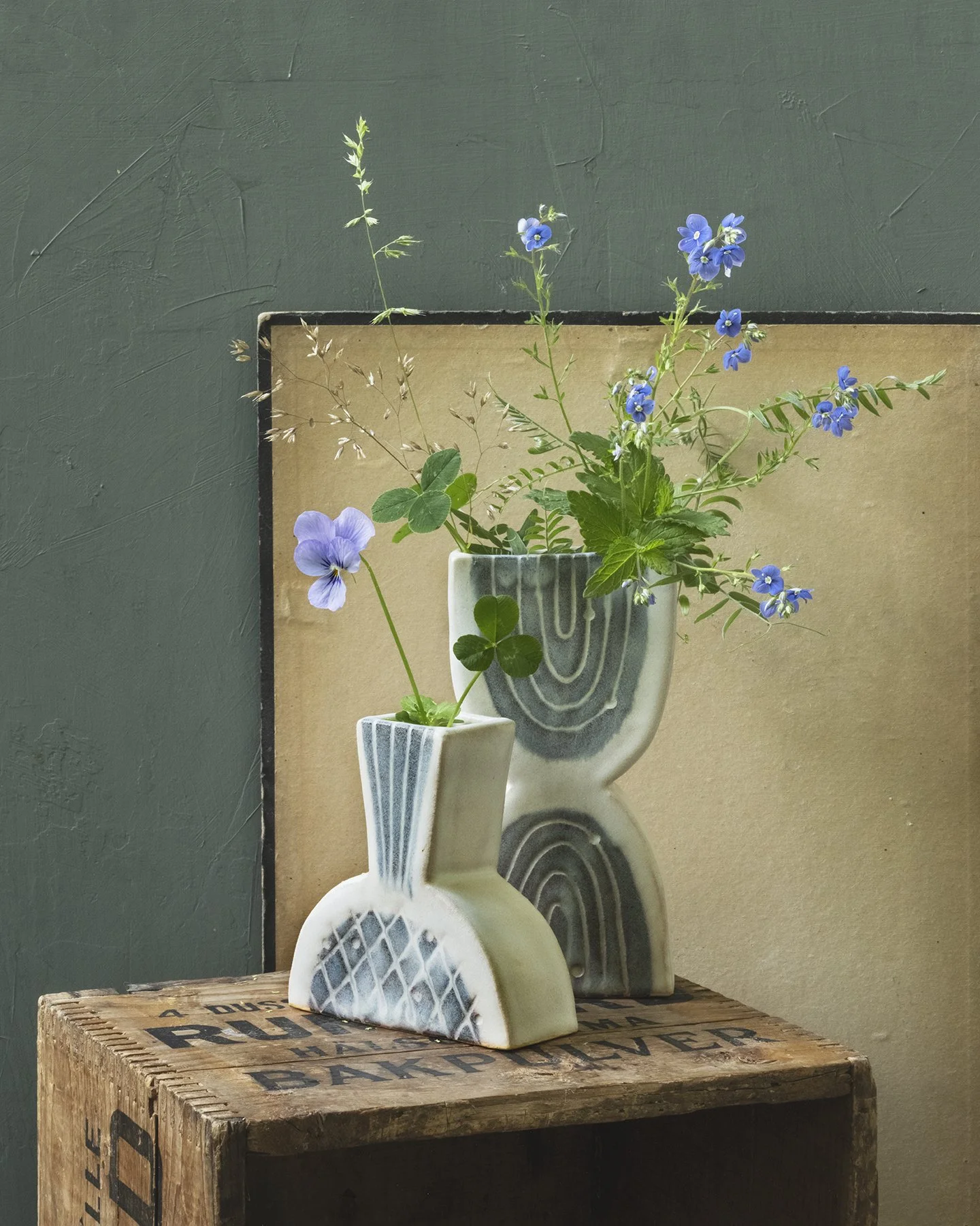 Two ceramic vases containing blue and purple flowers with green leaves, placed on a wooden surface against a dark green wall and a beige framed art piece.