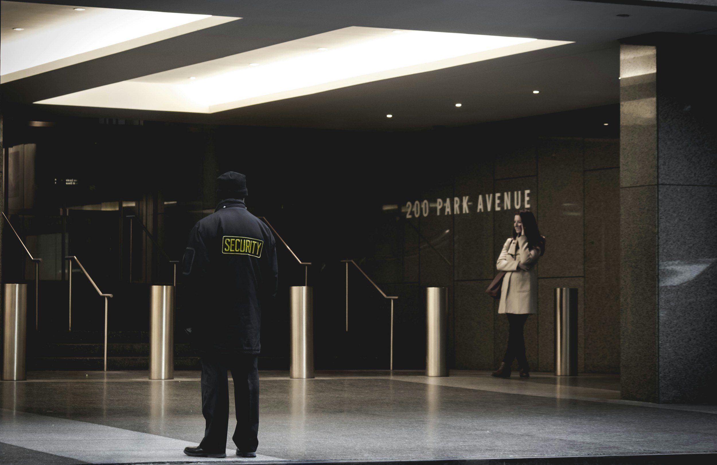 Security guard standing in front of a building entrance at 200 Park Avenue, with a woman standing near the wall, talking on her phone.