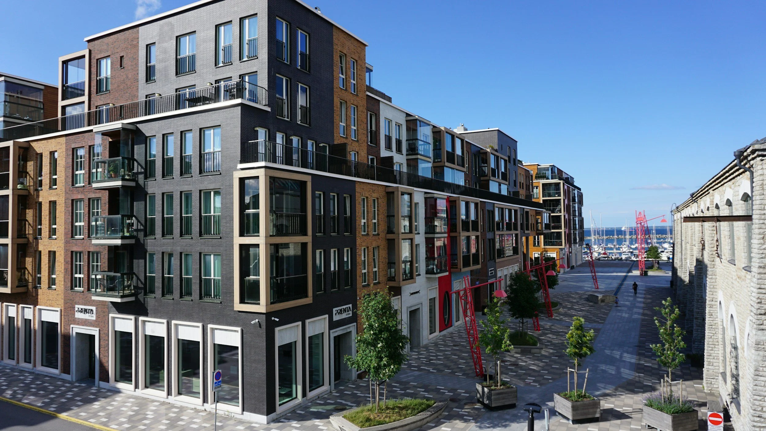 Modern multi-story residential building with large windows and balconies, adjacent to a pedestrian area with young trees, and a marina with sailboats in the background.